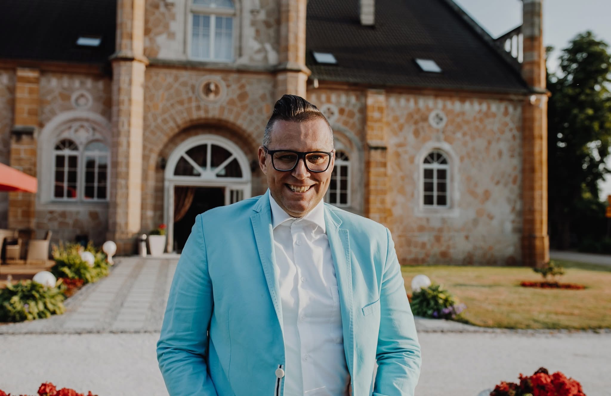 Uśmiechnięty mężczyzna w błękitnej marynarce na tle zabytkowego budynku z kamienia, prawdopodobnie podczas organizacji imprezy plenerowej.