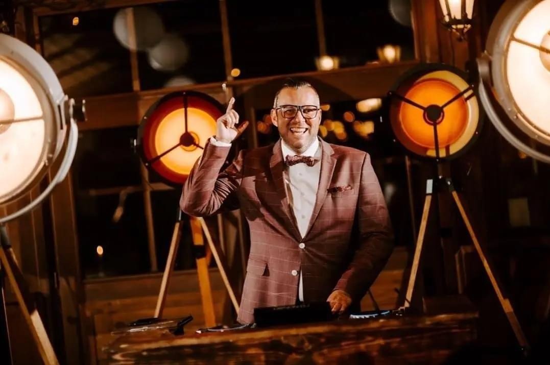 Uśmiechnięty DJ w bordowym garniturze i muszce, z uniesionym palcem, za konsoletą, otoczony przez reflektory w stylu retro, tworzące ciepłą atmosferę.