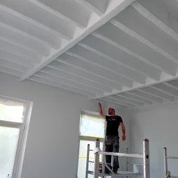 RM BAU Usługi Budowlane Robert Michalak - Mężczyzna na rusztowaniu maluje białą farbą sufit z drewnianymi belkami w jasnym wnętrzu. Okna zabezpieczone folią malarską, widoczne narzędzia pracy.