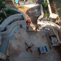 RM BAU Usługi Budowlane Robert Michalak - Budowa oczka wodnego z kamiennymi brzegami, widok z góry. W tle narzędzia i materiały budowlane. Prace w toku, widoczna siatka zbrojeniowa i częściowo wykończone brzegi.