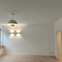 RM BAU Usługi Budowlane Robert Michalak - Jasne, parkietowe podłogi ułożone w jodełkę, białe ściany z nowoczesnym oświetleniem punktowym i wiszącą lampą. Widok na wykończone wnętrze z otwartymi drzwiami.