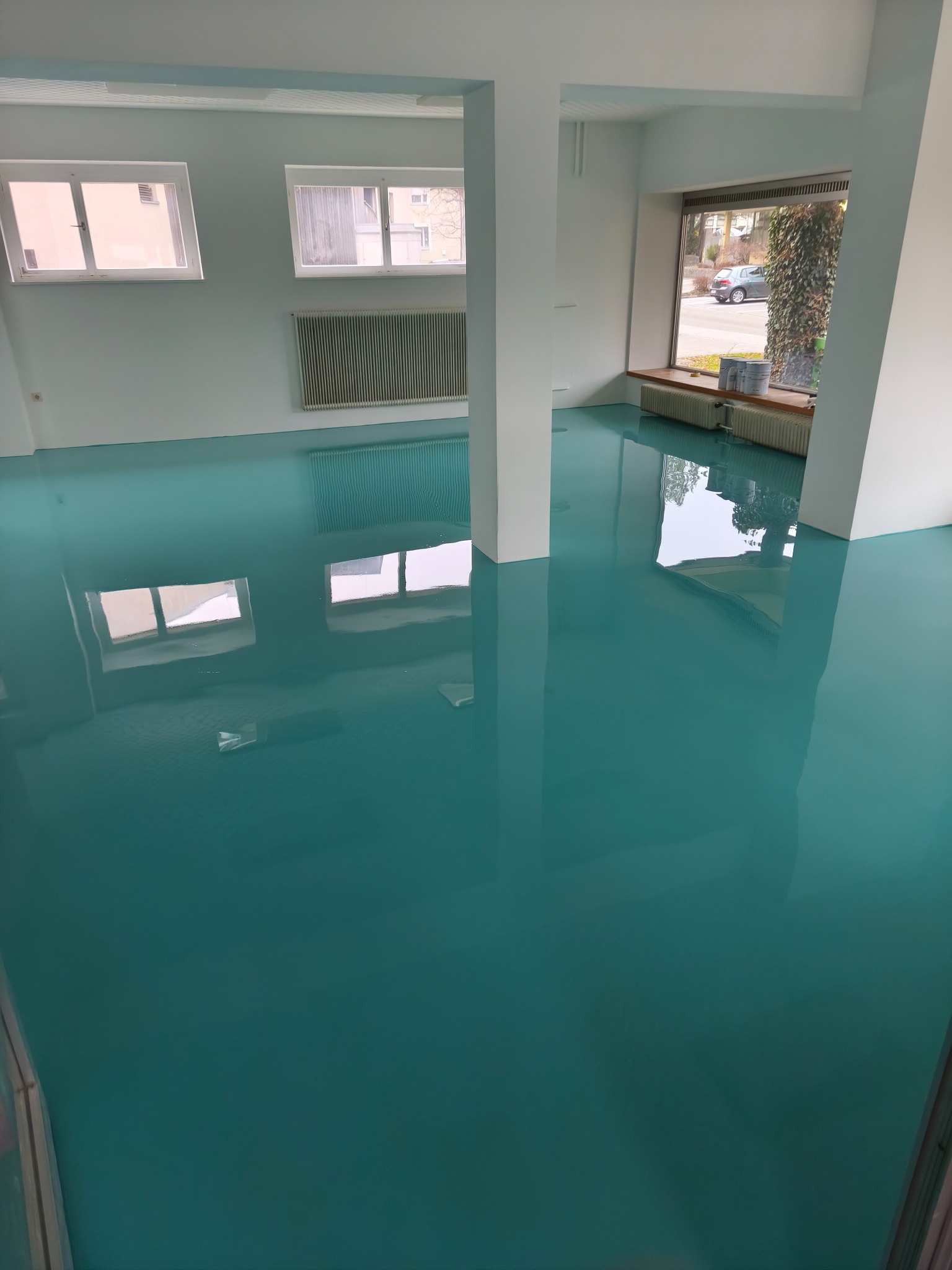 Błyszcząca, turkusowa podłoga w jasnym, minimalistycznym wnętrzu z białymi ścianami i kolumnami. Okna odbijają światło, tworząc lustrzany efekt na podłodze.