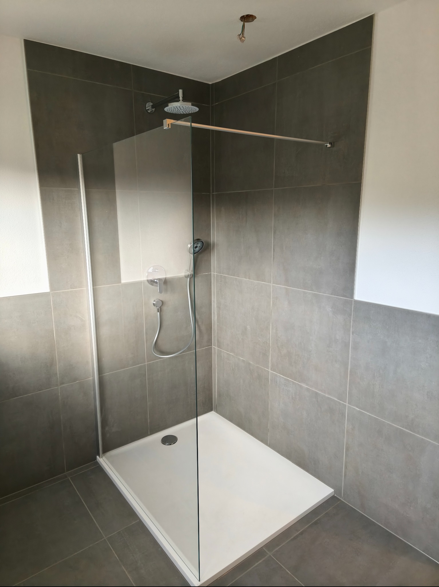 Nowoczesna kabina prysznicowa z szarymi płytkami i białą brodzikiem. Minimalistyczny design, szklana ścianka, chromowana armatura. Widoczne łączenia płytek.