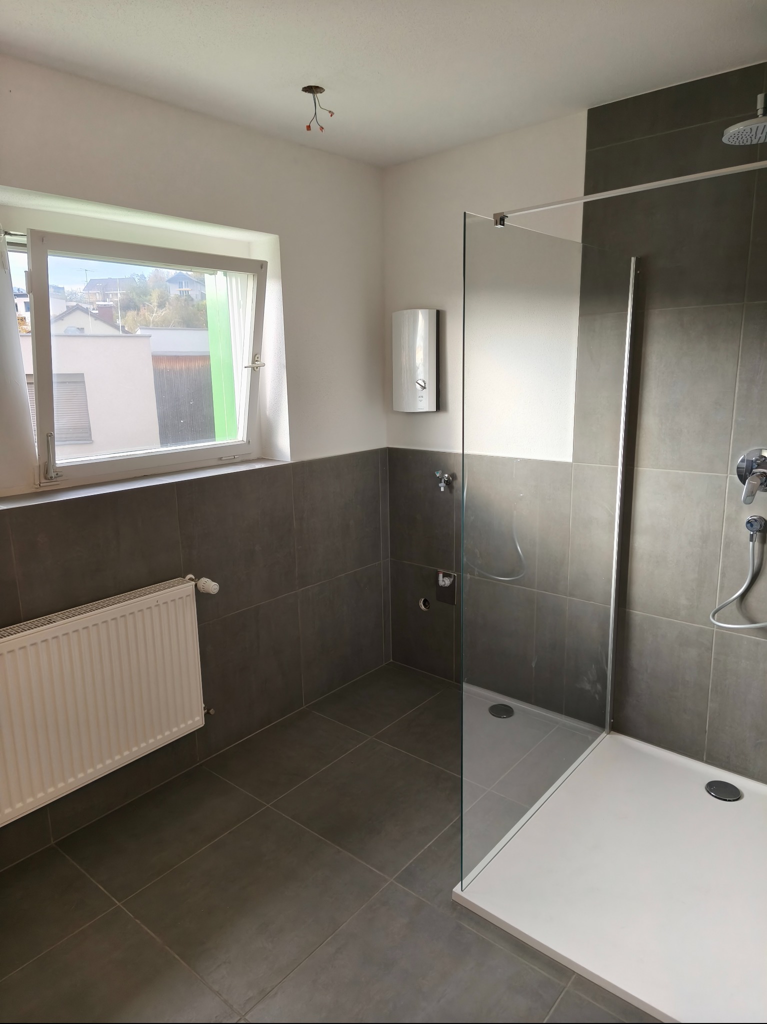 Nowoczesna łazienka z szarymi płytkami, kabiną prysznicową typu walk-in i białym grzejnikiem pod oknem. Widoczne przyłącza elektryczne na suficie.