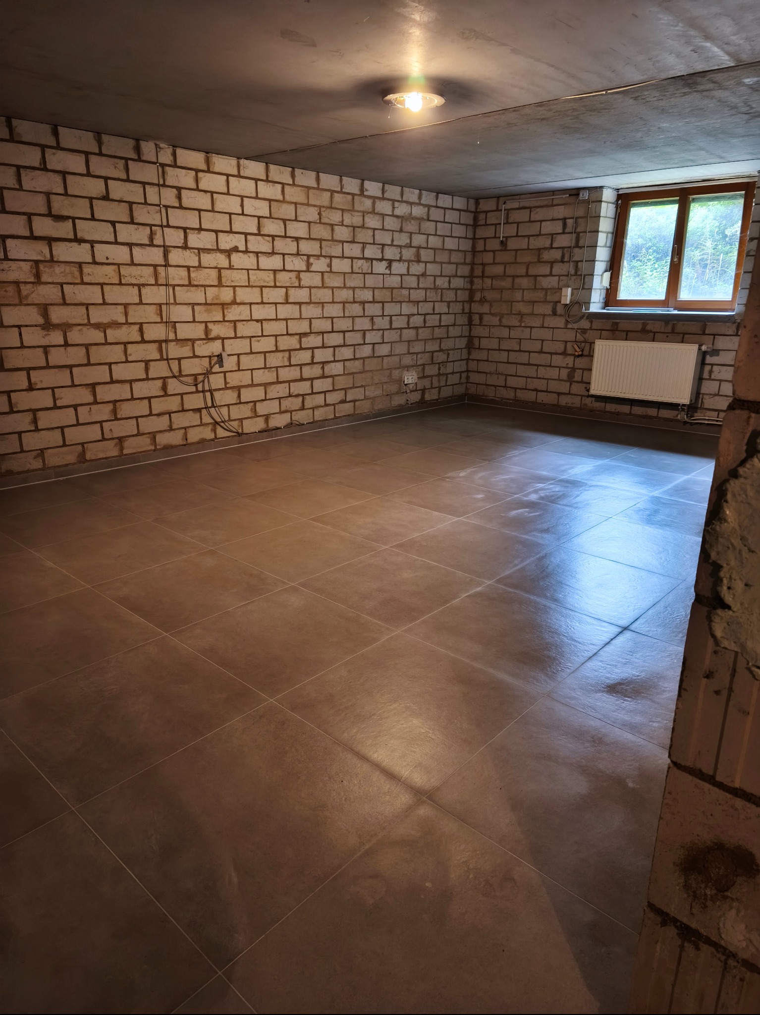 Wykończone wnętrze piwnicy z brązowymi płytkami podłogowymi, surowymi ścianami z cegły i oknem z widokiem na zieleń. Widoczny grzejnik i surowy betonowy sufit.