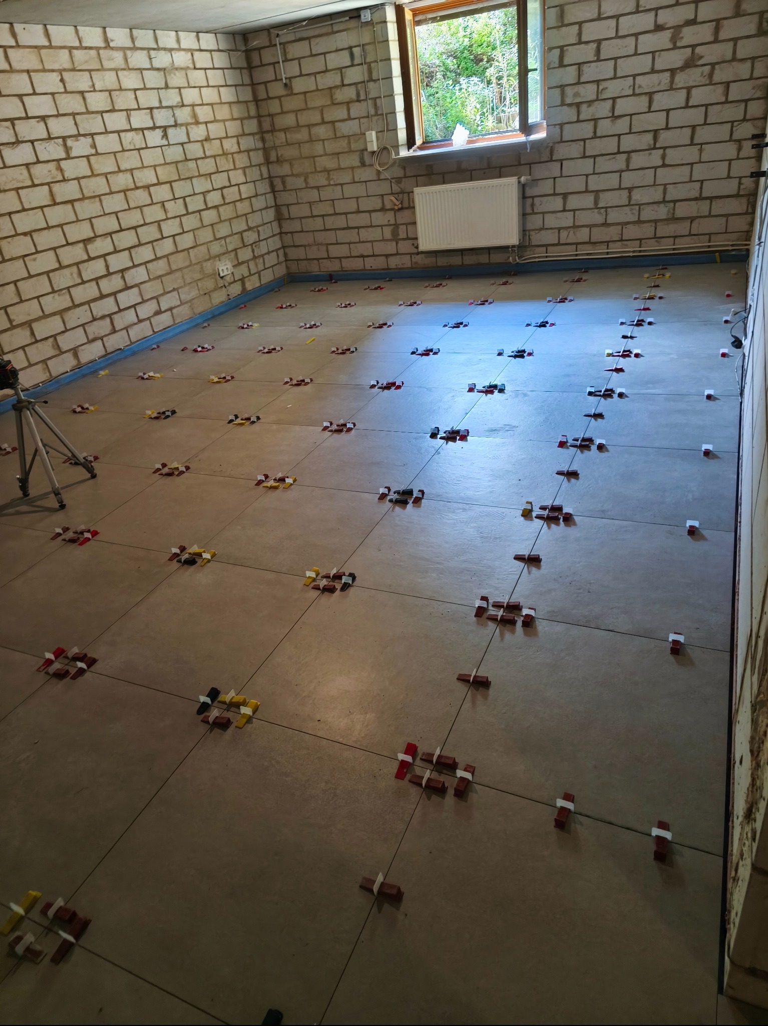 Ułożone płytki podłogowe w pomieszczeniu z surowymi ścianami z cegły, z użyciem systemu poziomowania płytek. Widoczne kolorowe kliny i krzyżyki dystansowe.