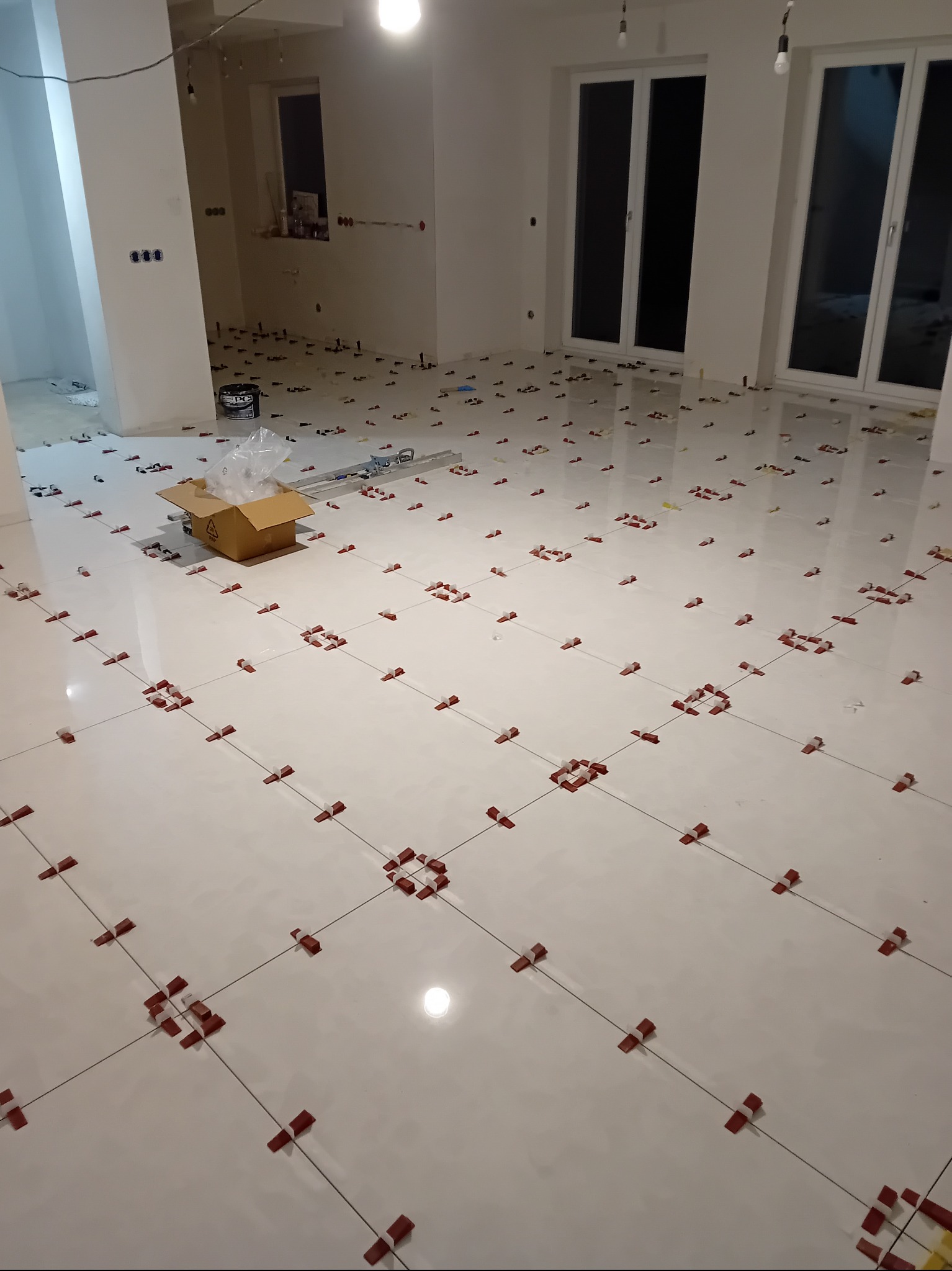 Białe płytki podłogowe w trakcie układania, z czerwonymi klipsami poziomującymi i krzyżykami dystansowymi. W tle widoczne okna i przygotowane ściany.
