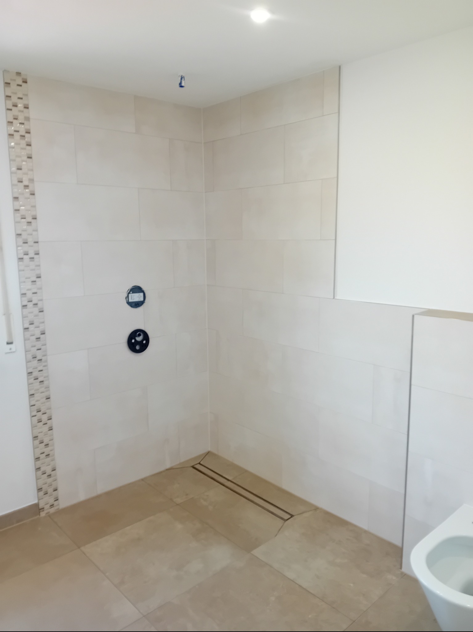 Jasna łazienka po remoncie: beżowe płytki na ścianach i podłodze, dekoracyjny pas mozaiki, odpływ liniowy w kabinie prysznicowej, przygotowane miejsce na armaturę.