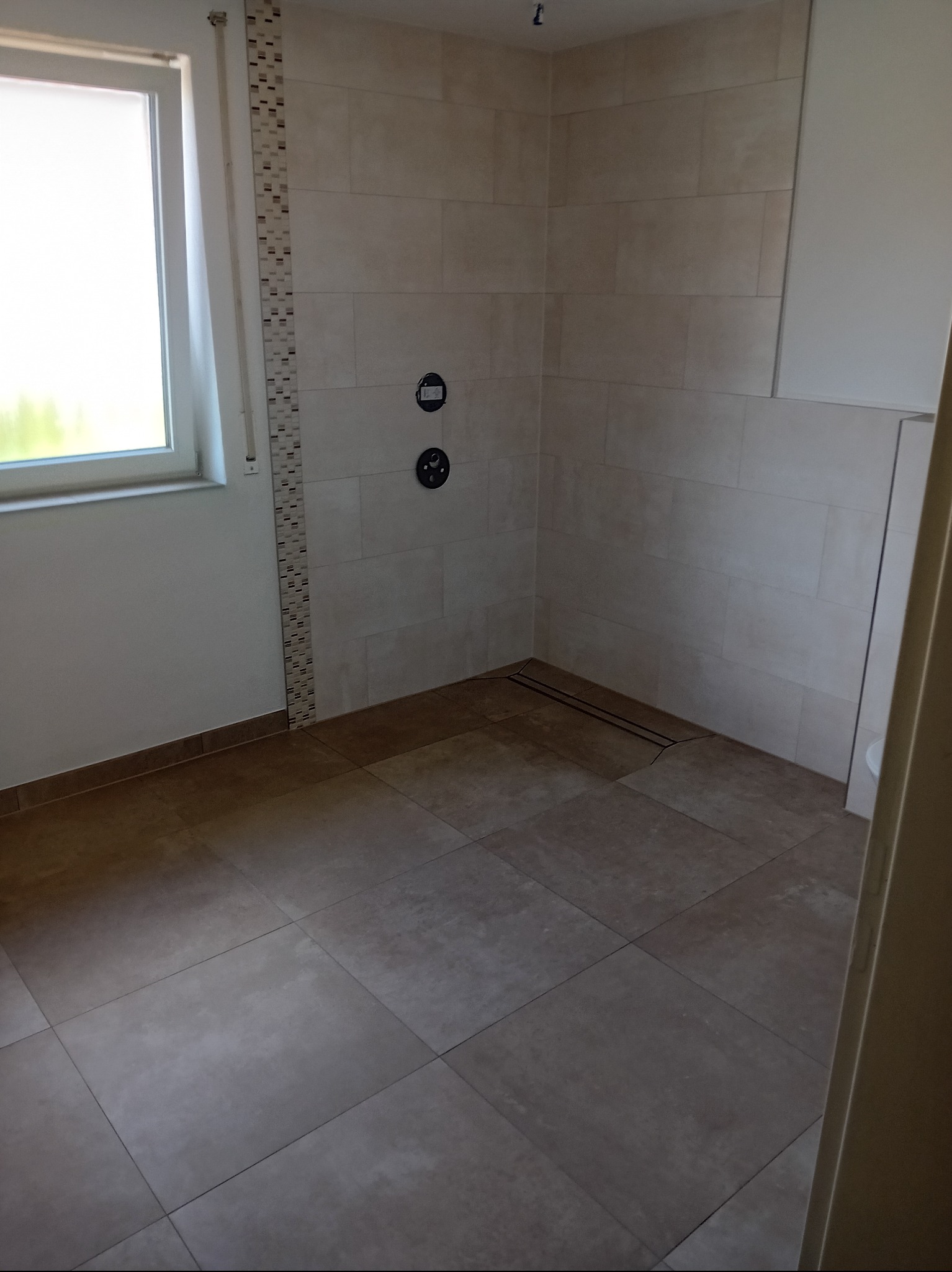 Beżowa łazienka z oknem i mozaiką. Widoczne płytki na ścianach i podłodze, armatura oraz odpływ liniowy. Minimalistyczny design, jasne wnętrze.