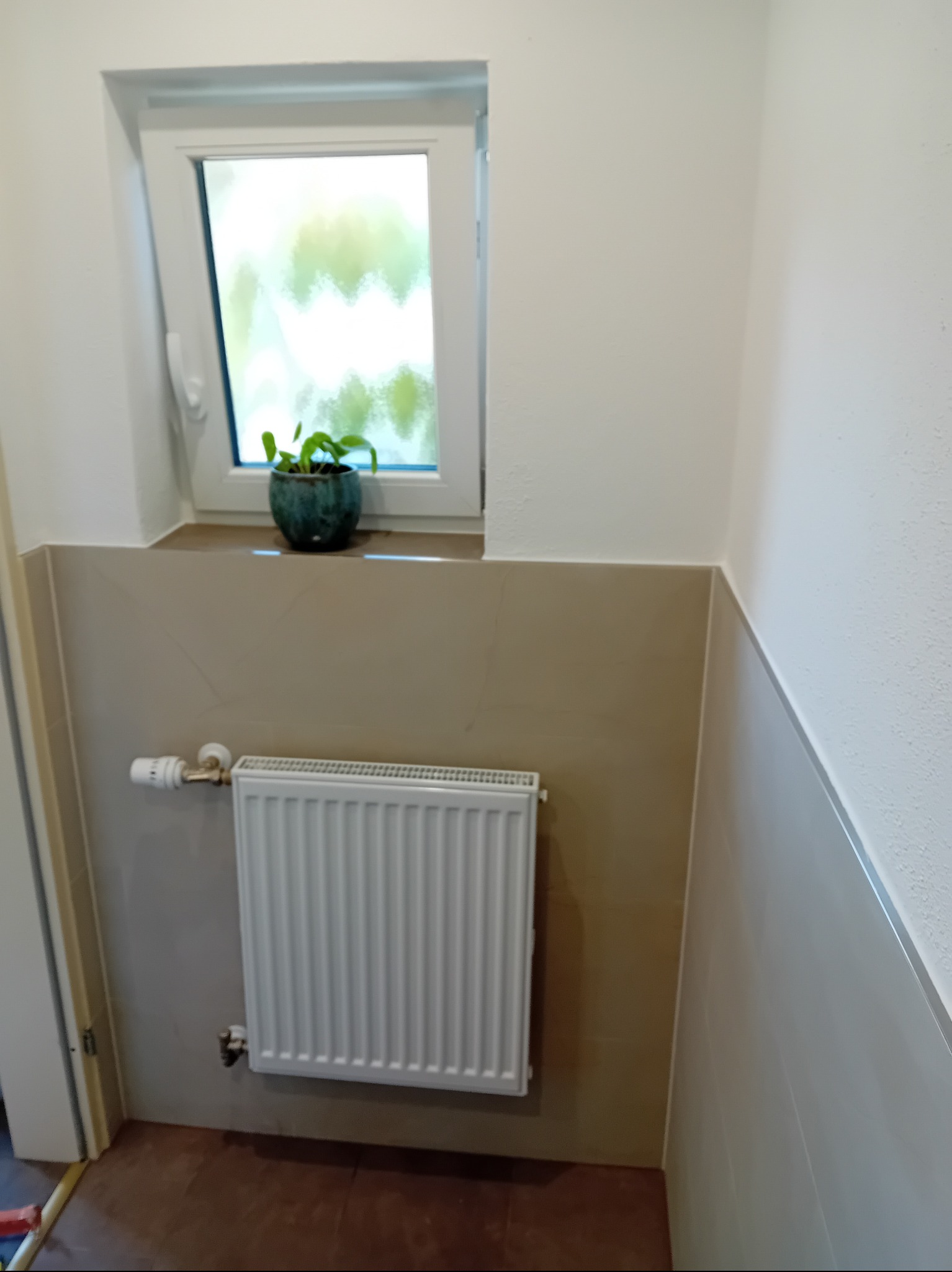 Wykończona łazienka z grzejnikiem, brązowymi płytkami do połowy ściany i białymi ścianami powyżej. Na parapecie okna stoi doniczka z zieloną rośliną.