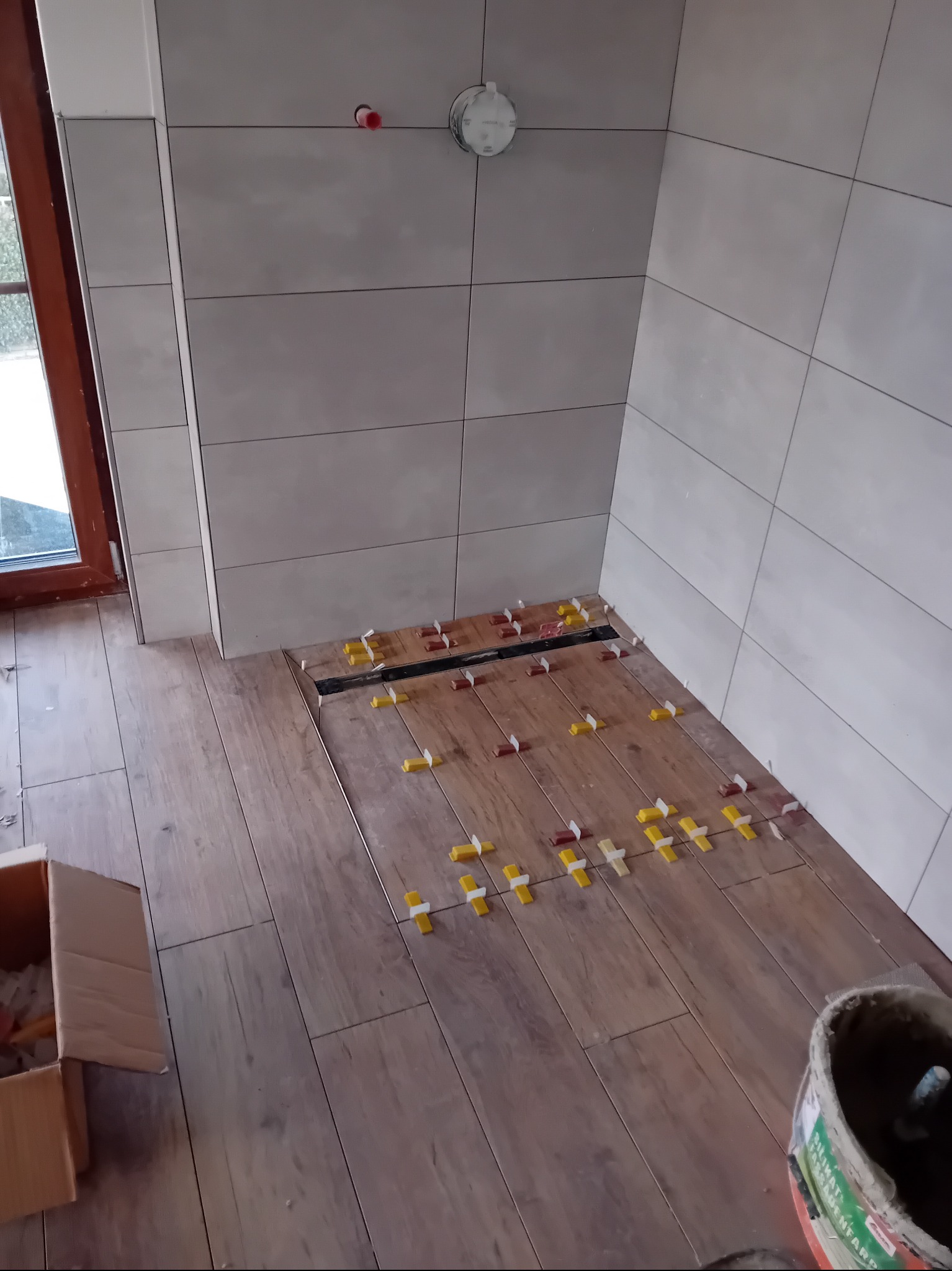 Układanie drewnopodobnych płytek podłogowych w łazience z odpływem liniowym. Na ścianach jasne, kwadratowe płytki. Widoczne krzyżyki dystansowe.