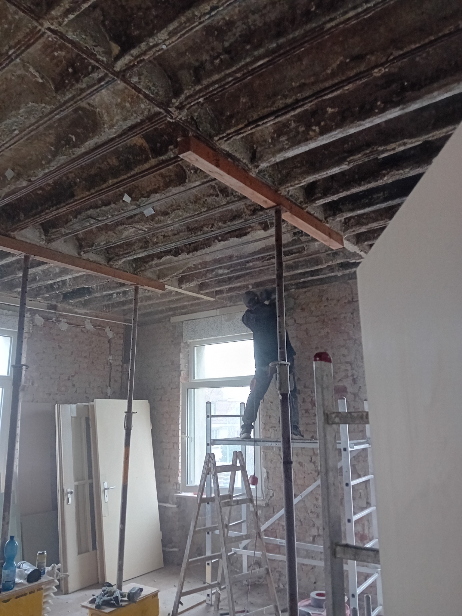 Pracownik na rusztowaniu montuje drewniane belki pod sufitem w remontowanym pomieszczeniu z odsłoniętą cegłą i widoczną konstrukcją stropu.
