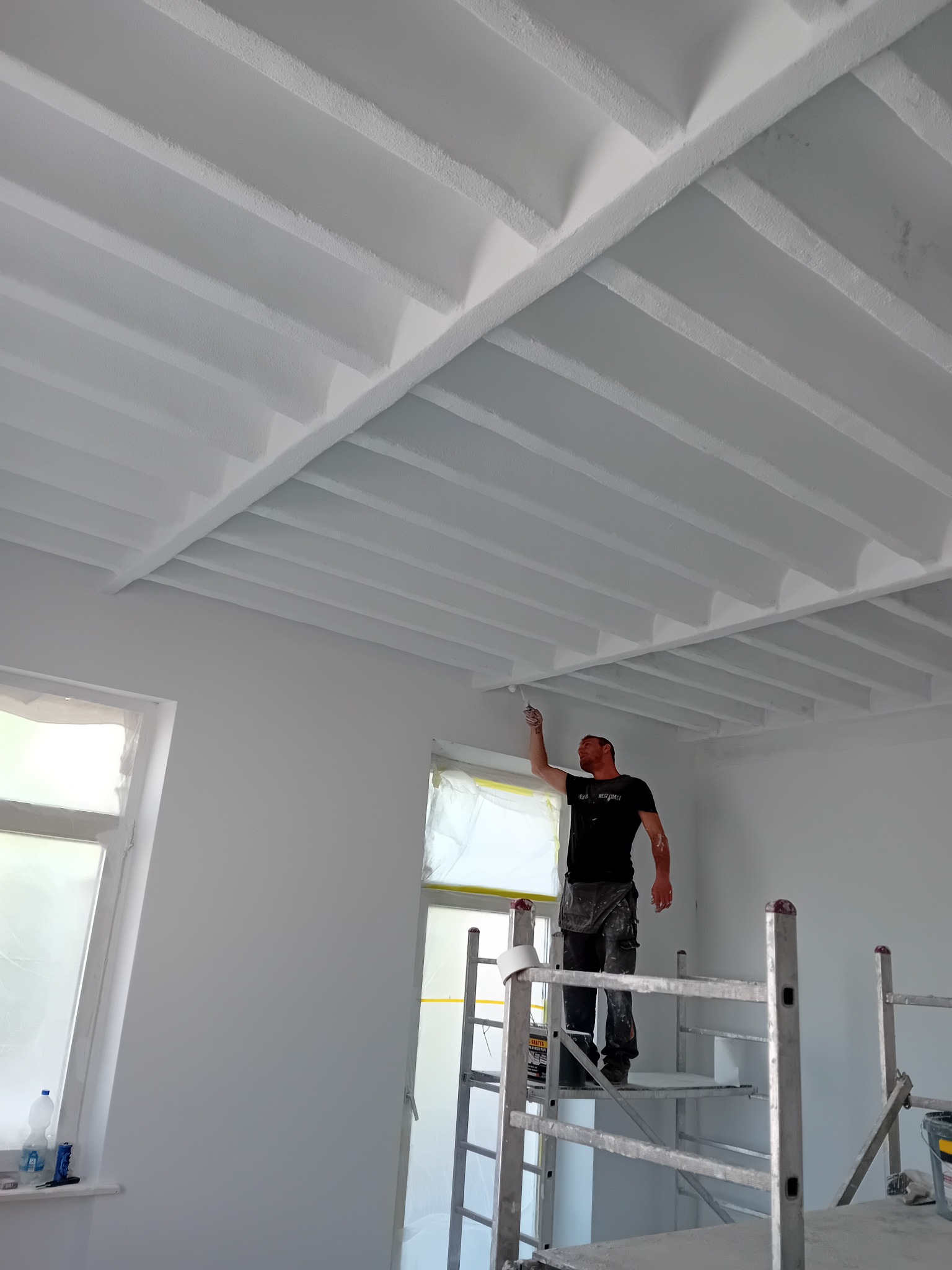 Mężczyzna na rusztowaniu maluje białą farbą sufit z drewnianymi belkami w jasnym wnętrzu. Okna zabezpieczone folią malarską, widoczne narzędzia pracy.