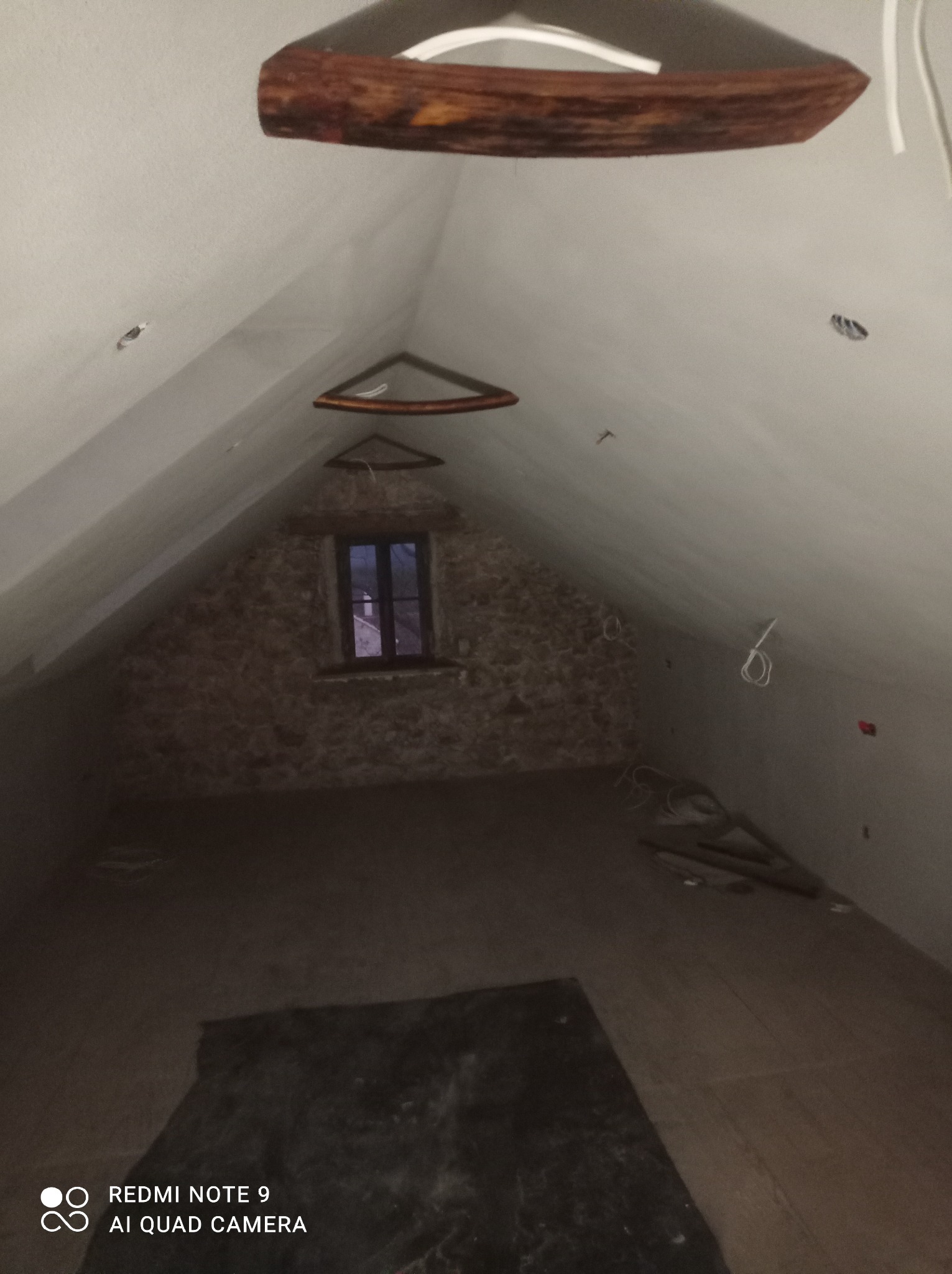 Poddasze po remoncie: białe ściany, drewniane elementy dekoracyjne, okno w kamiennej ścianie. Widoczne kable elektryczne, podłoga częściowo zakryta folią.