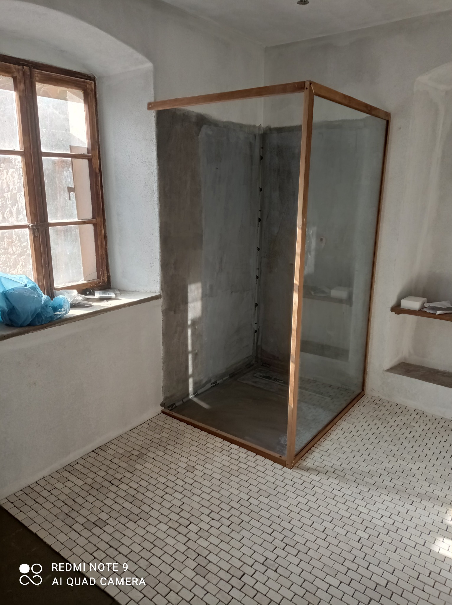 Surowa kabina prysznicowa z drewnianą ramą i szklanymi ścianami w łazience z mozaikową podłogą i oknem. Wnętrze w trakcie wykończenia, widoczne ślady prac.