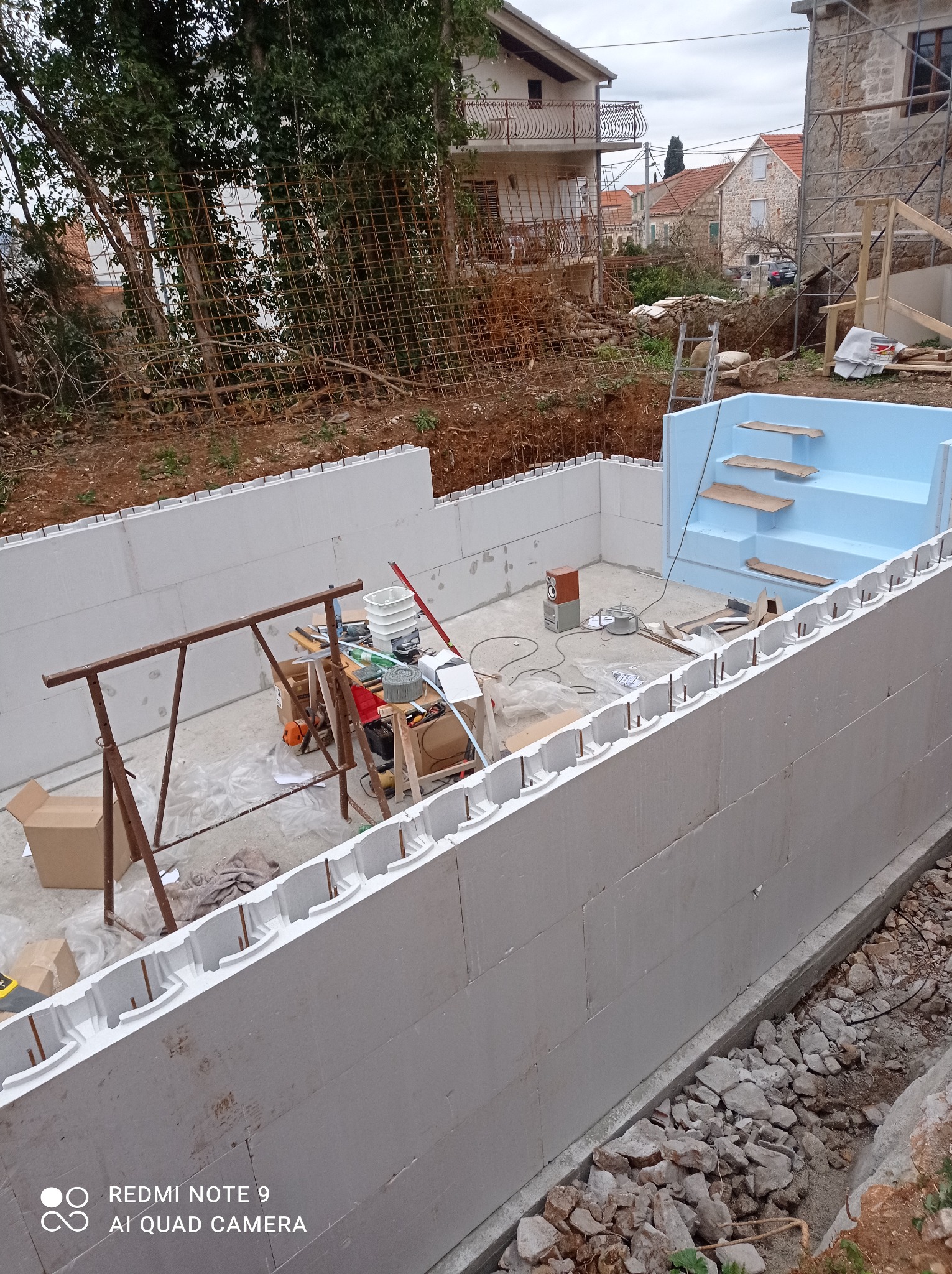 Budowa basenu: białe ściany z pustaków szalunkowych, niebieska niecka schodkowa, narzędzia i materiały budowlane na placu budowy w ogrodzie.