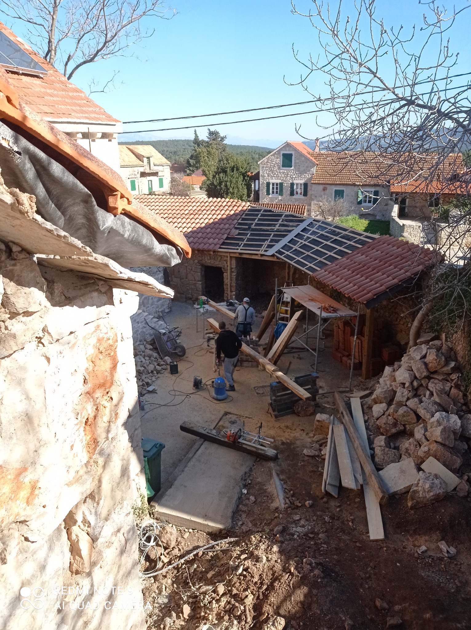 Budowa kamiennego budynku z czerwoną dachówką w słoneczny dzień. Widoczne rusztowania, materiały budowlane i pracownicy w trakcie pracy.