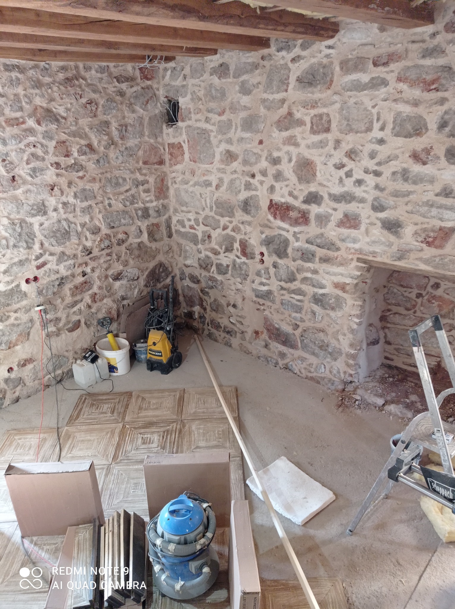 Wnętrze w trakcie prac wykończeniowych: surowe kamienne ściany, częściowo ułożona podłoga z płytek, narzędzia i materiały budowlane. Widoczny odkurzacz przemysłowy i drabina.
