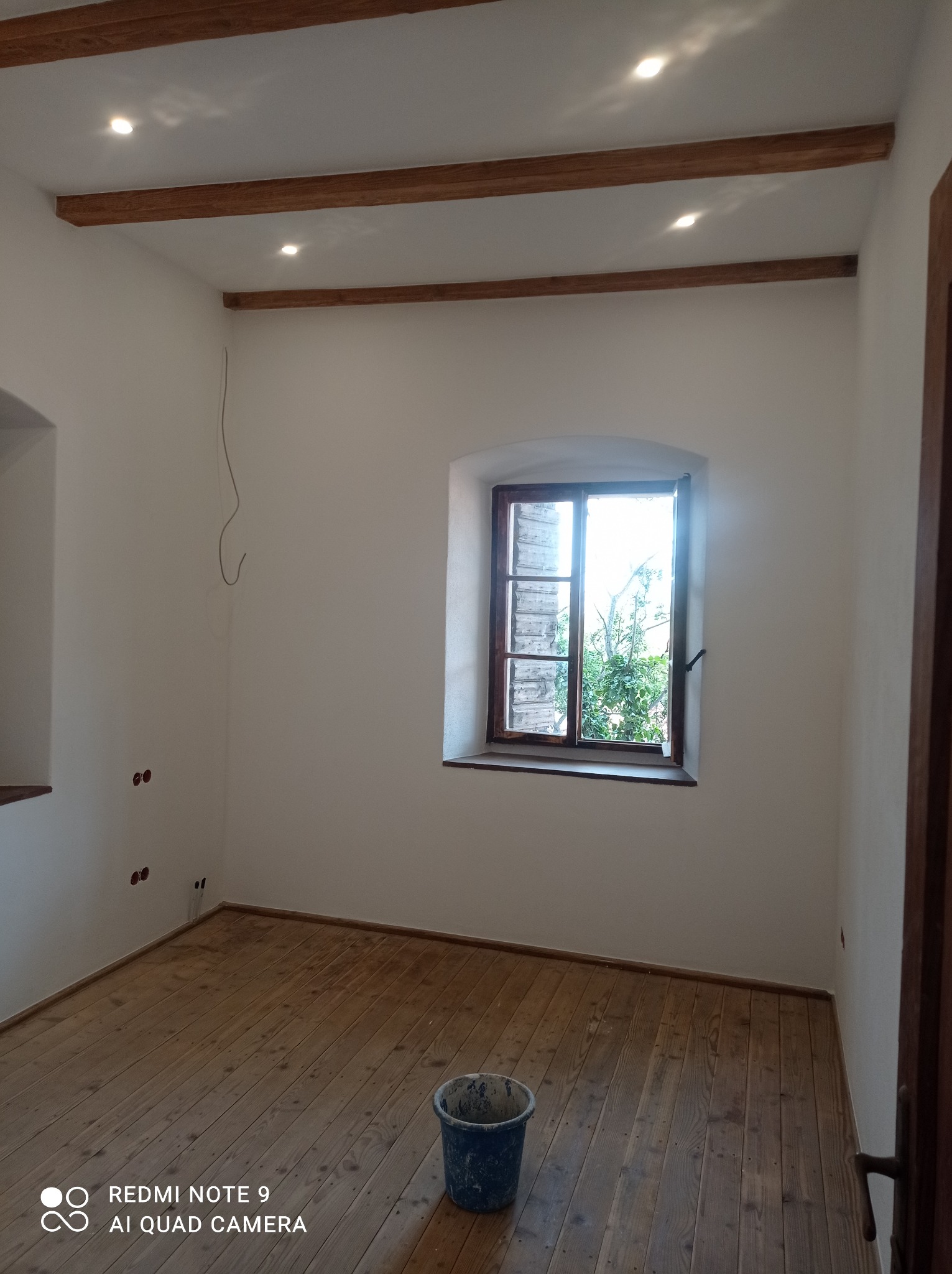 Wnętrze po remoncie: białe ściany, drewniana podłoga i belki stropowe, okno z widokiem na zieleń. Wiadro z farbą w rogu. Widoczne gniazdka elektryczne.