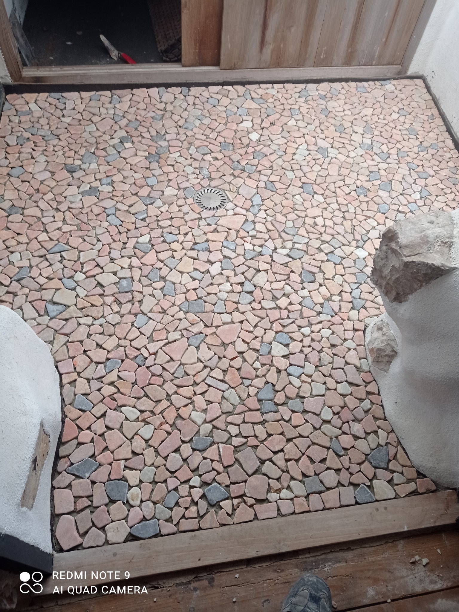 Mozaika z nieregularnych kamieni w odcieniach różu i szarości, tworząca unikalną posadzkę w łazience z odpływem liniowym. Widoczne fragmenty ścian i drewniane wykończenie.