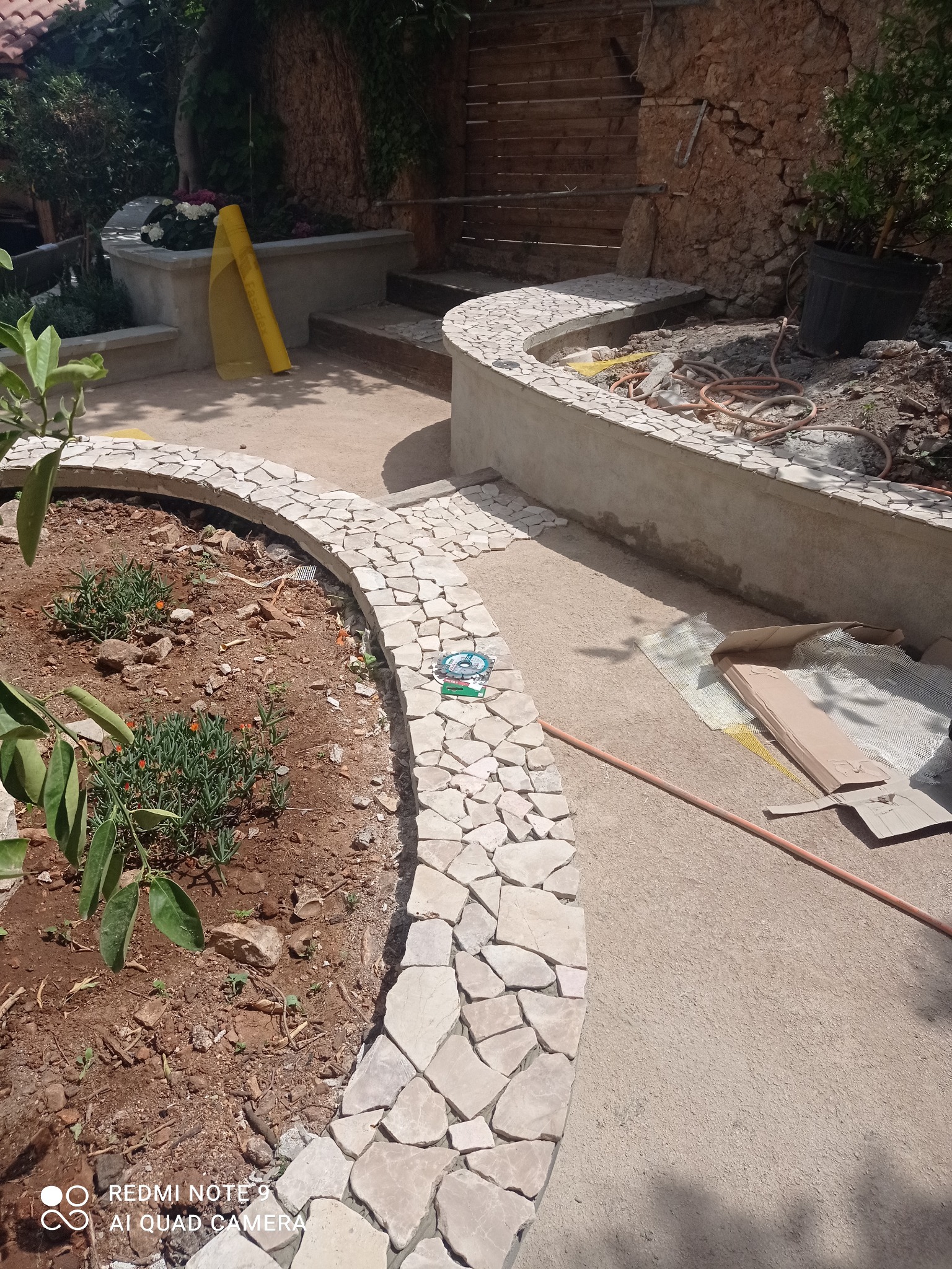 Kaskadowy ogród z kamiennymi obrzeżami rabat i ścieżką w trakcie budowy. Widoczne narzędzia i materiały budowlane. Jasne, naturalne światło.