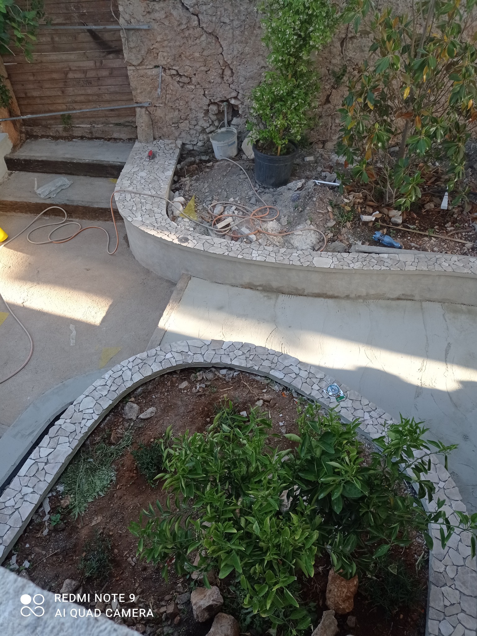 Kaskadowy ogród w trakcie budowy: betonowe obrzeża wykończone mozaiką z kamienia naturalnego, rośliny w ziemi, narzędzia budowlane w tle, wysoka perspektywa.