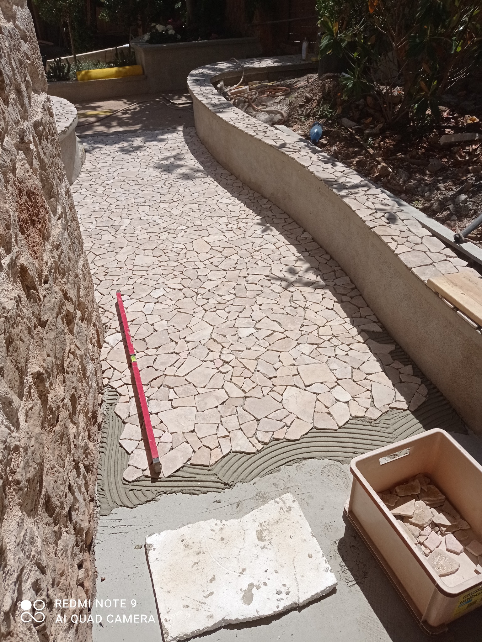 Układanie mozaiki z nieregularnych płytek na ścieżce. Widoczny poziomica, pojemnik z płytkami i świeży klej. Fragment ścieżki już wykończony.