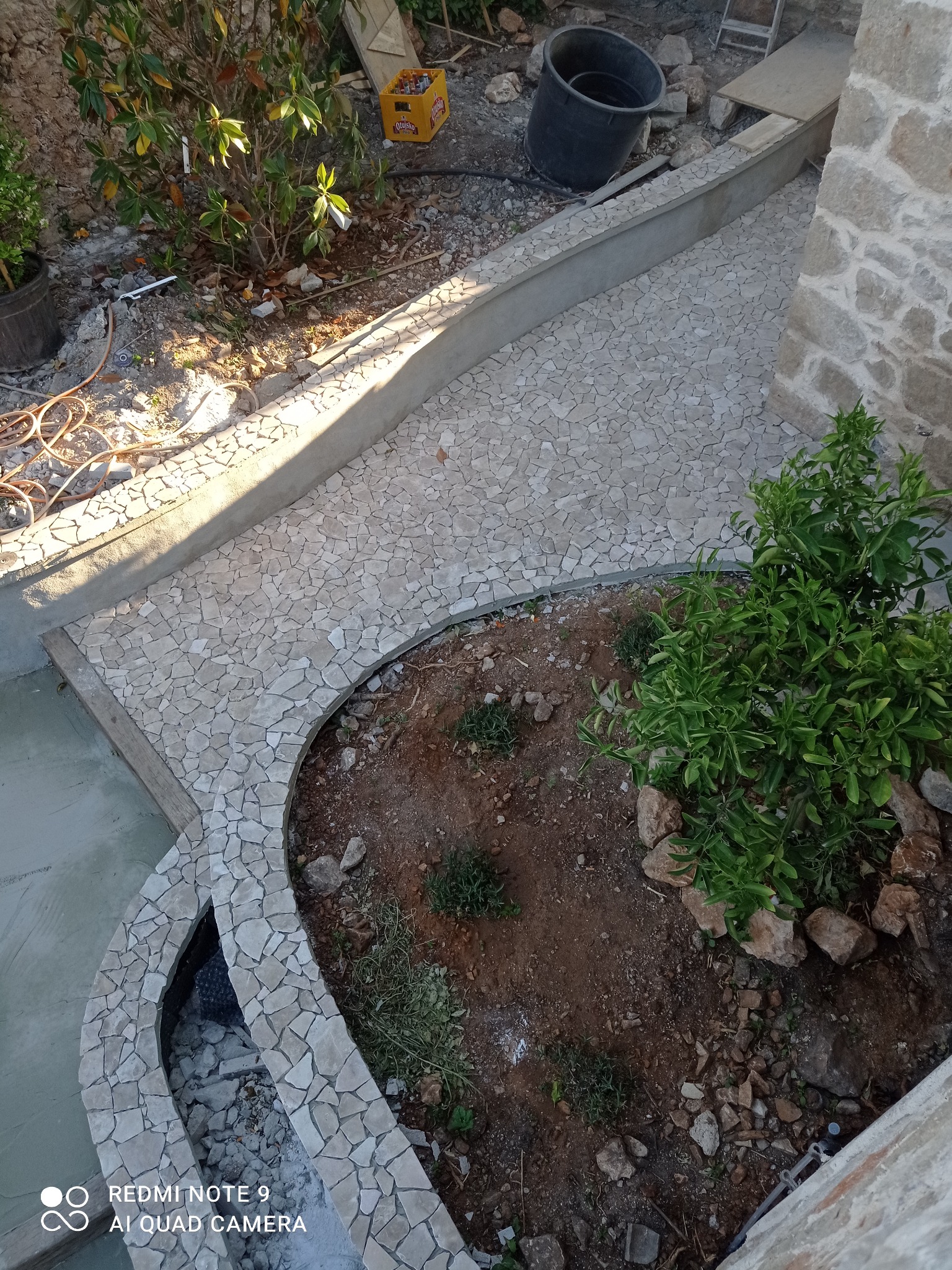 Ułożona mozaika z kamienia naturalnego tworząca ścieżkę i obrzeże rabaty w ogrodzie. Widok z góry, jasne kamienie, roślinność. Detal wykończenia ogrodu.