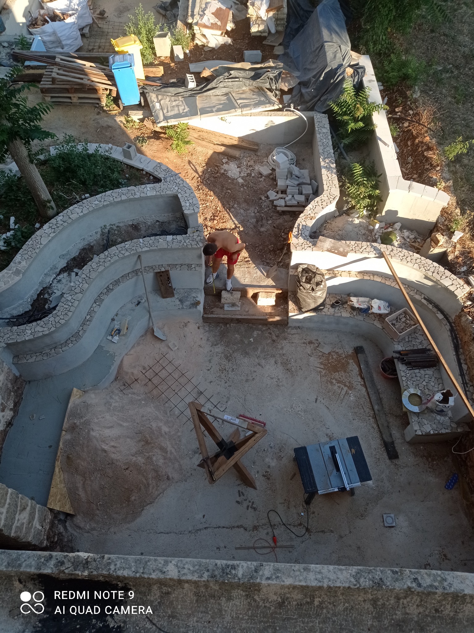 Budowa oczka wodnego z kamiennymi brzegami, widok z góry. W tle narzędzia i materiały budowlane. Prace w toku, widoczna siatka zbrojeniowa i częściowo wykończone brzegi.