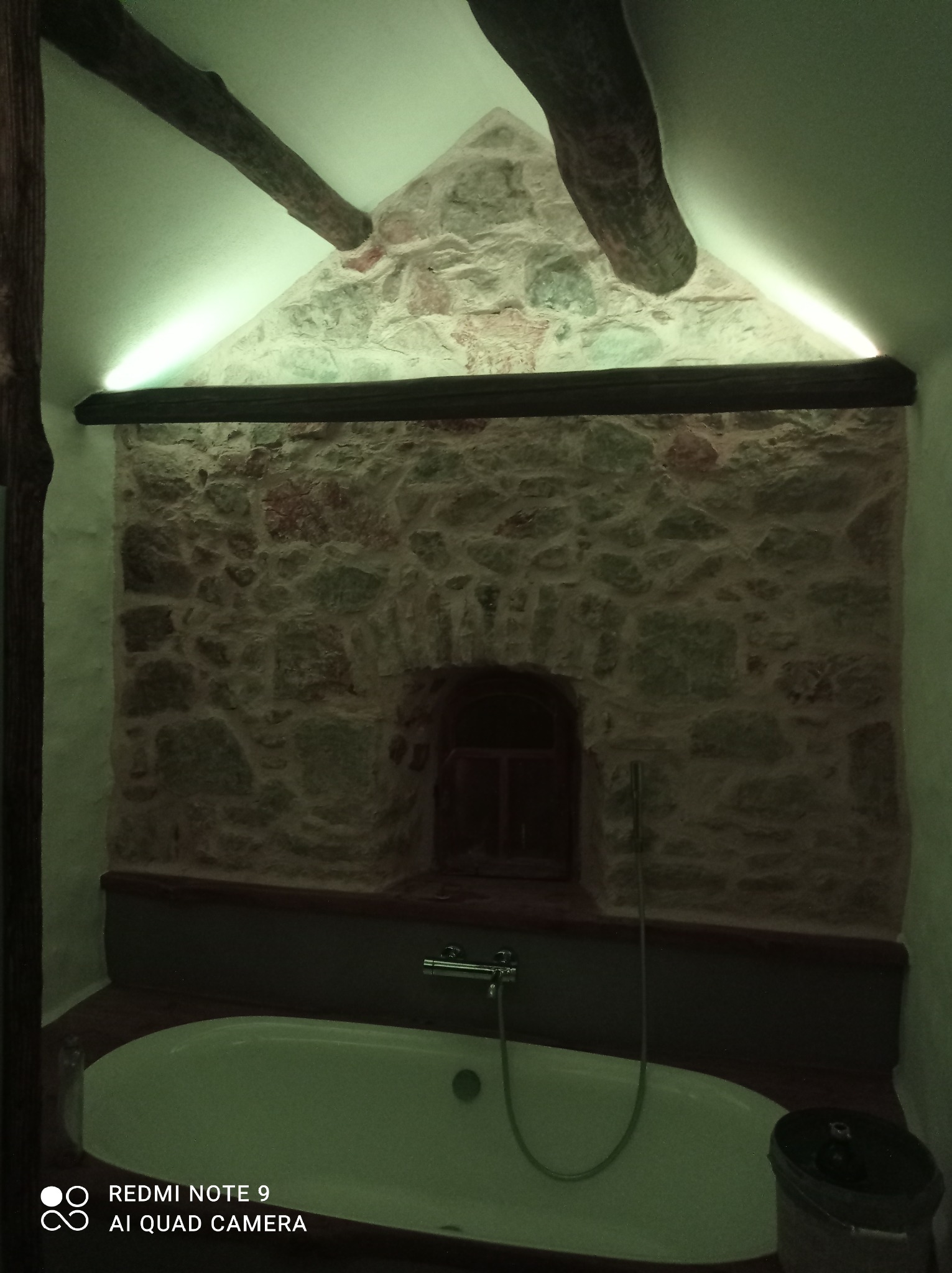 Rustykalna łazienka z kamienną ścianą, drewnianymi belkami i podświetleniem LED nad wanną. W tle małe okienko. Całość utrzymana w stonowanej kolorystyce.