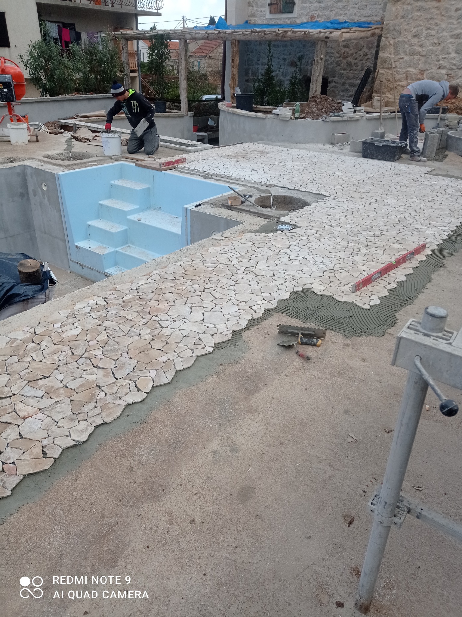 Układanie mozaiki z kamienia naturalnego wokół basenu. Dwóch pracowników w trakcie pracy, widoczne narzędzia i poziomica. Jasne płytki basenowe.