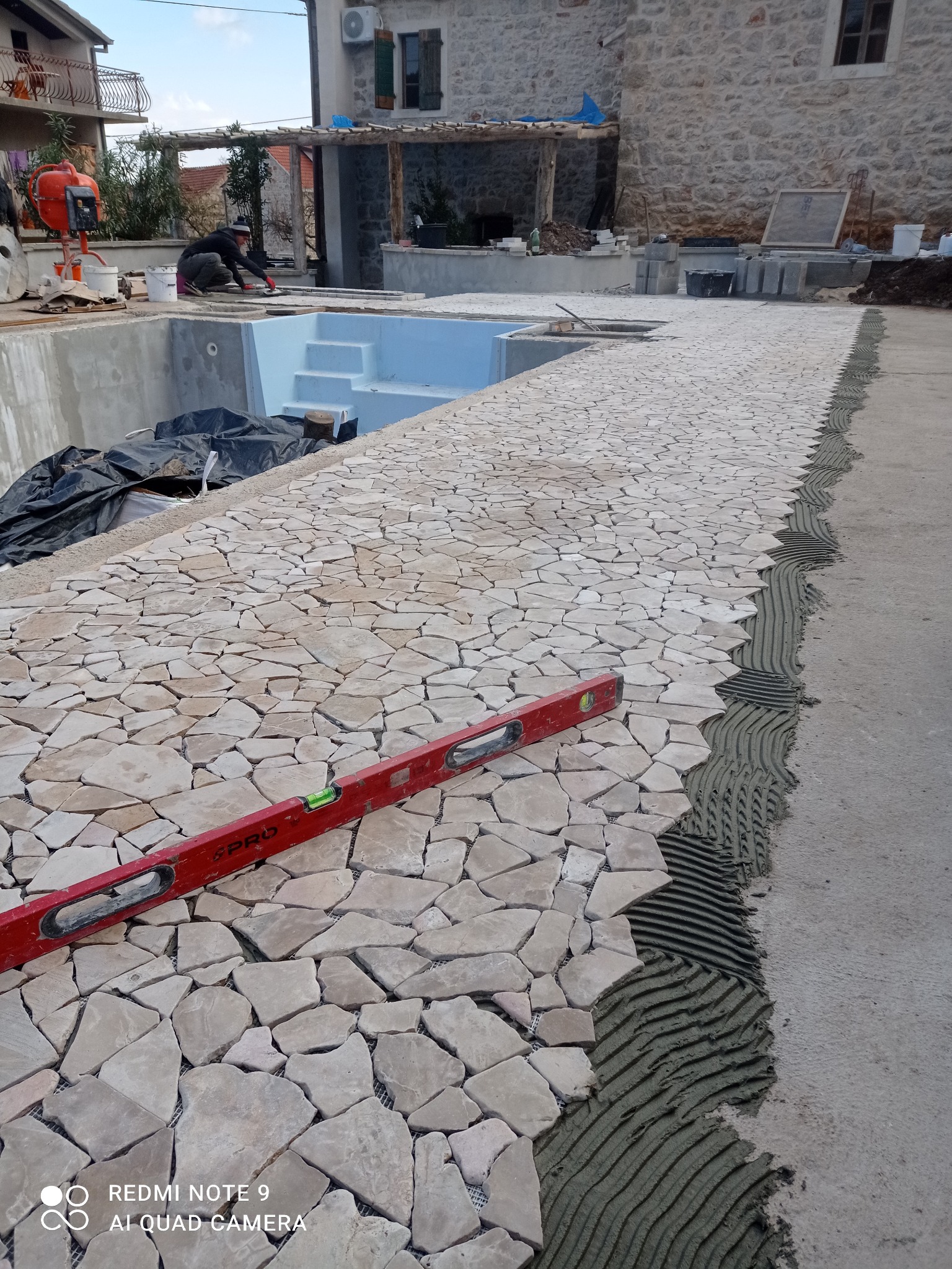 Układanie mozaiki z nieregularnych kamieni wokół basenu, widoczny poziomica i świeżo nałożony klej. W tle dom z kamienia i pracownik przy betoniarce.