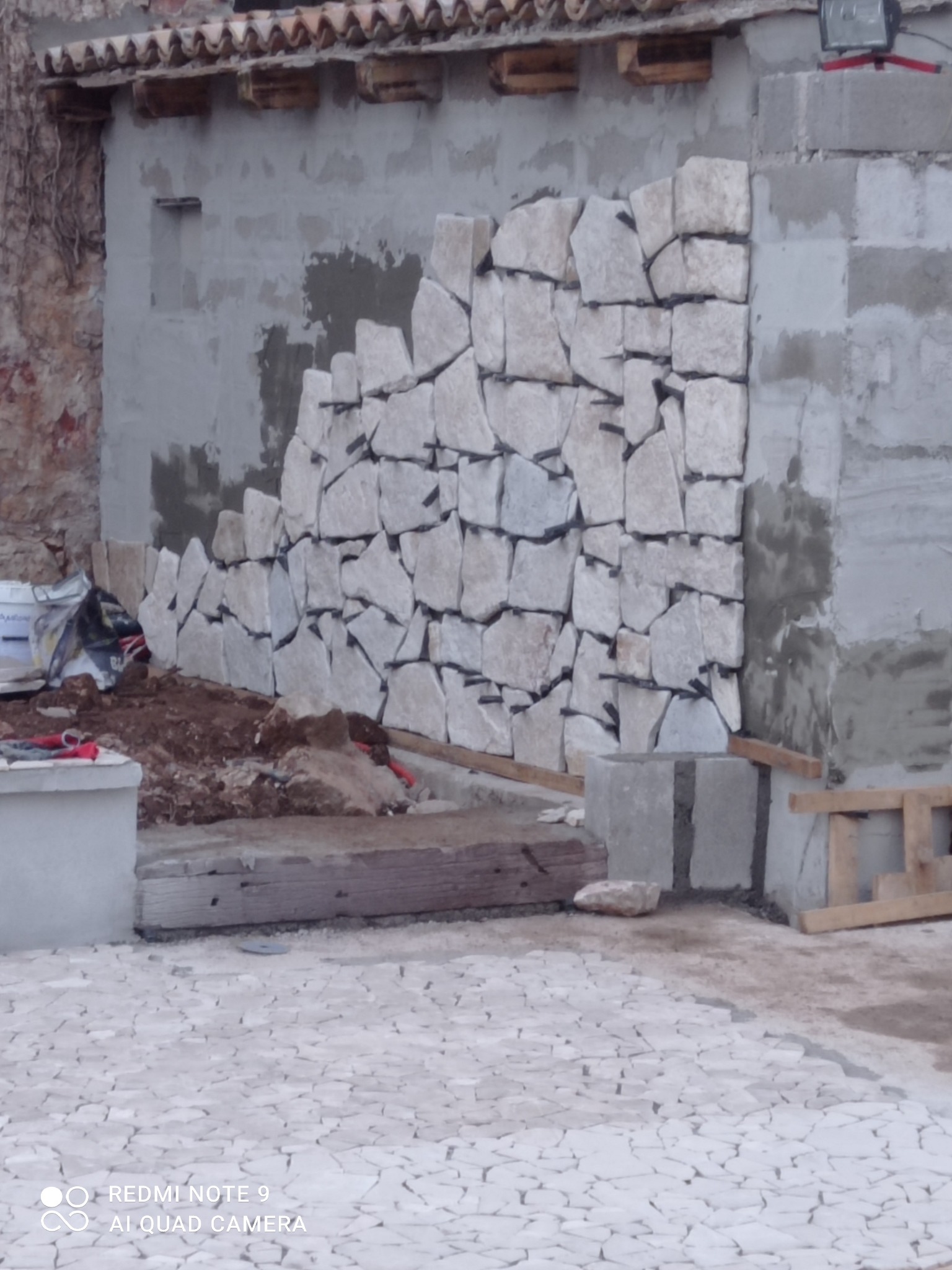 Ściana budynku w trakcie wykańczania kamieniem naturalnym o nieregularnych kształtach, widoczne narzędzia i materiały budowlane. Prace wykończeniowe w toku.