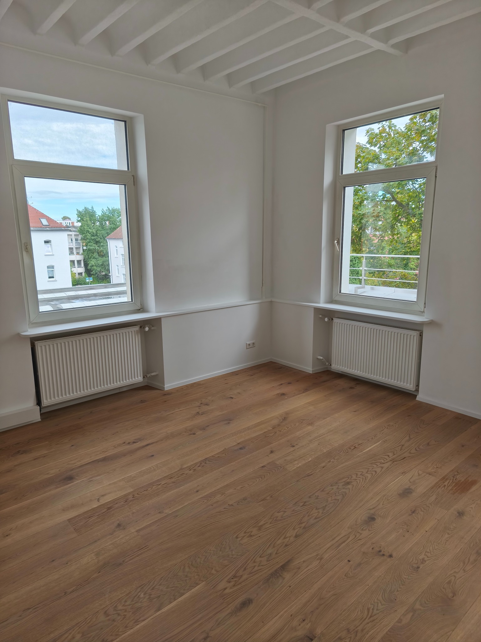 Jasne, puste wnętrze z drewnianą podłogą, białymi ścianami i dwoma oknami z widokiem na zieleń. Grzejniki pod oknami. Sufit z dekoracyjnymi belkami.