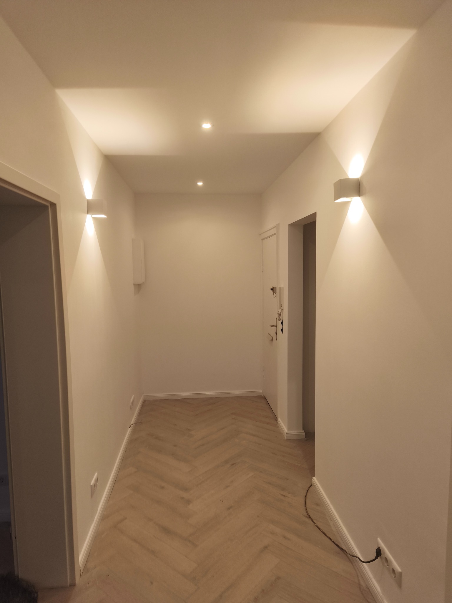 Korytarz po remoncie: parkiet w jodełkę, białe ściany z nowoczesnymi kinkietami i punktowym oświetleniem sufitowym. Widoczne gniazdka elektryczne przy podłodze.