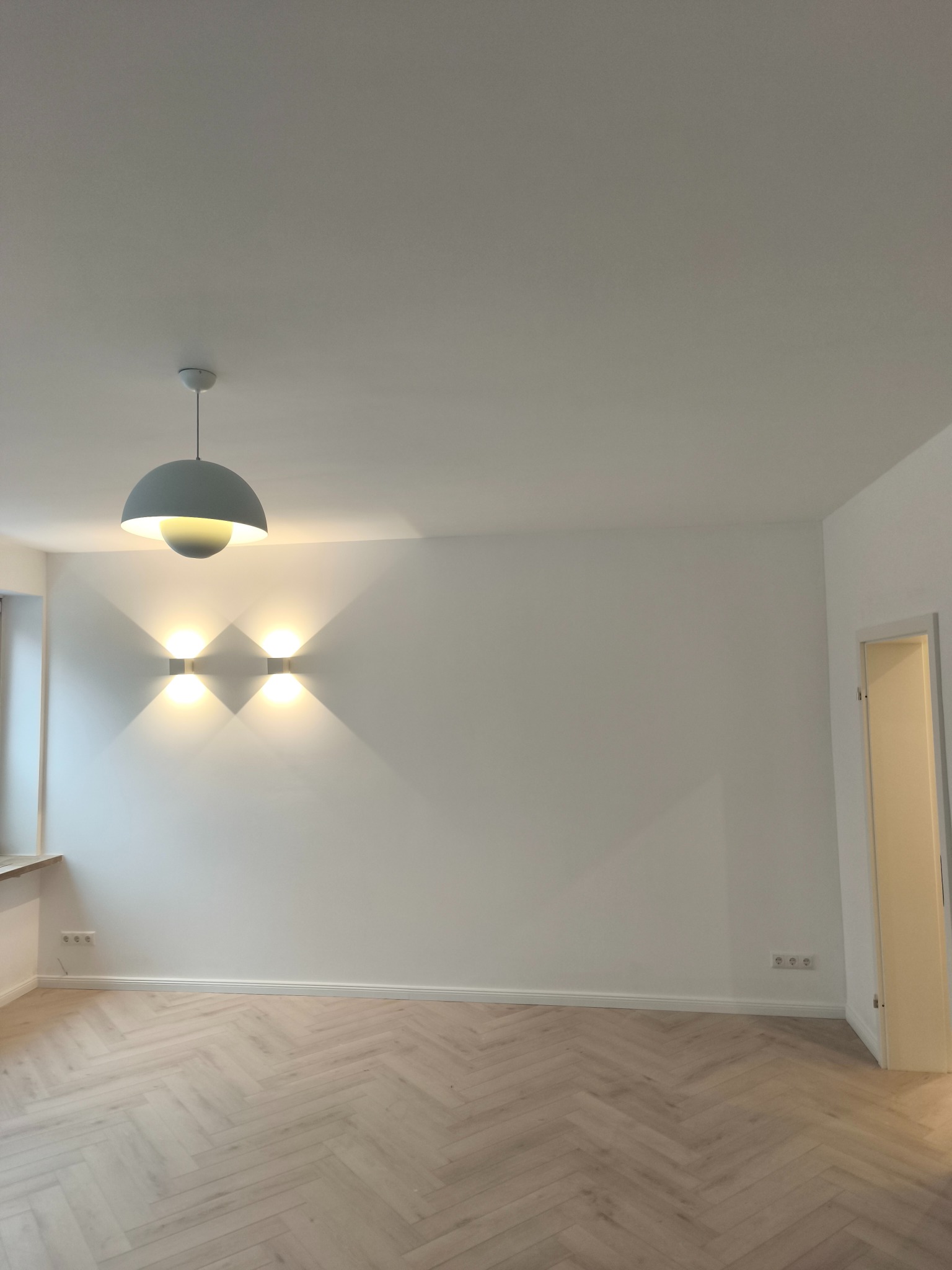 Jasne, parkietowe podłogi ułożone w jodełkę, białe ściany z nowoczesnym oświetleniem punktowym i wiszącą lampą. Widok na wykończone wnętrze z otwartymi drzwiami.