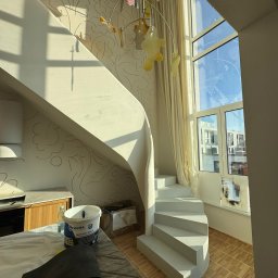 A.P. Kompleks - Jasne wnętrze z spiralnymi schodami po malowaniu, widoczne narzędzia malarskie i wiadro z farbą Sigma. Ściany z delikatnym wzorem, duże okno z widokiem na miasto.