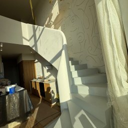 A.P. Kompleks - Białe schody w jasnym wnętrzu z dekoracyjną ścianą i zasłoniętym oknem. Widoczny fragment kuchni i lampa wisząca. Świeże malowanie, wpadające promienie słoneczne.