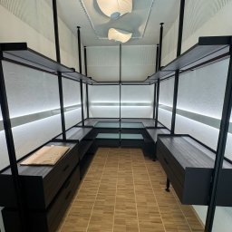 A.P. Kompleks - Nowoczesna garderoba z czarnymi regałami i szufladami, oświetleniem LED i beżową podłogą. Na półce leży brązowa paczka. Minimalistyczny design.