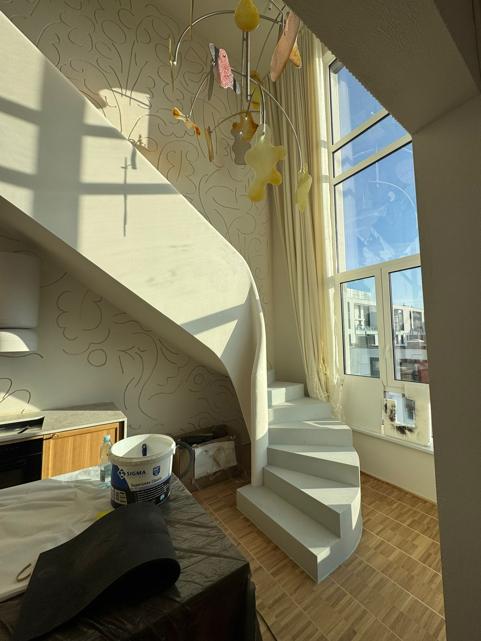 Jasne wnętrze z spiralnymi schodami po malowaniu, widoczne narzędzia malarskie i wiadro z farbą Sigma. Ściany z delikatnym wzorem, duże okno z widokiem na miasto.