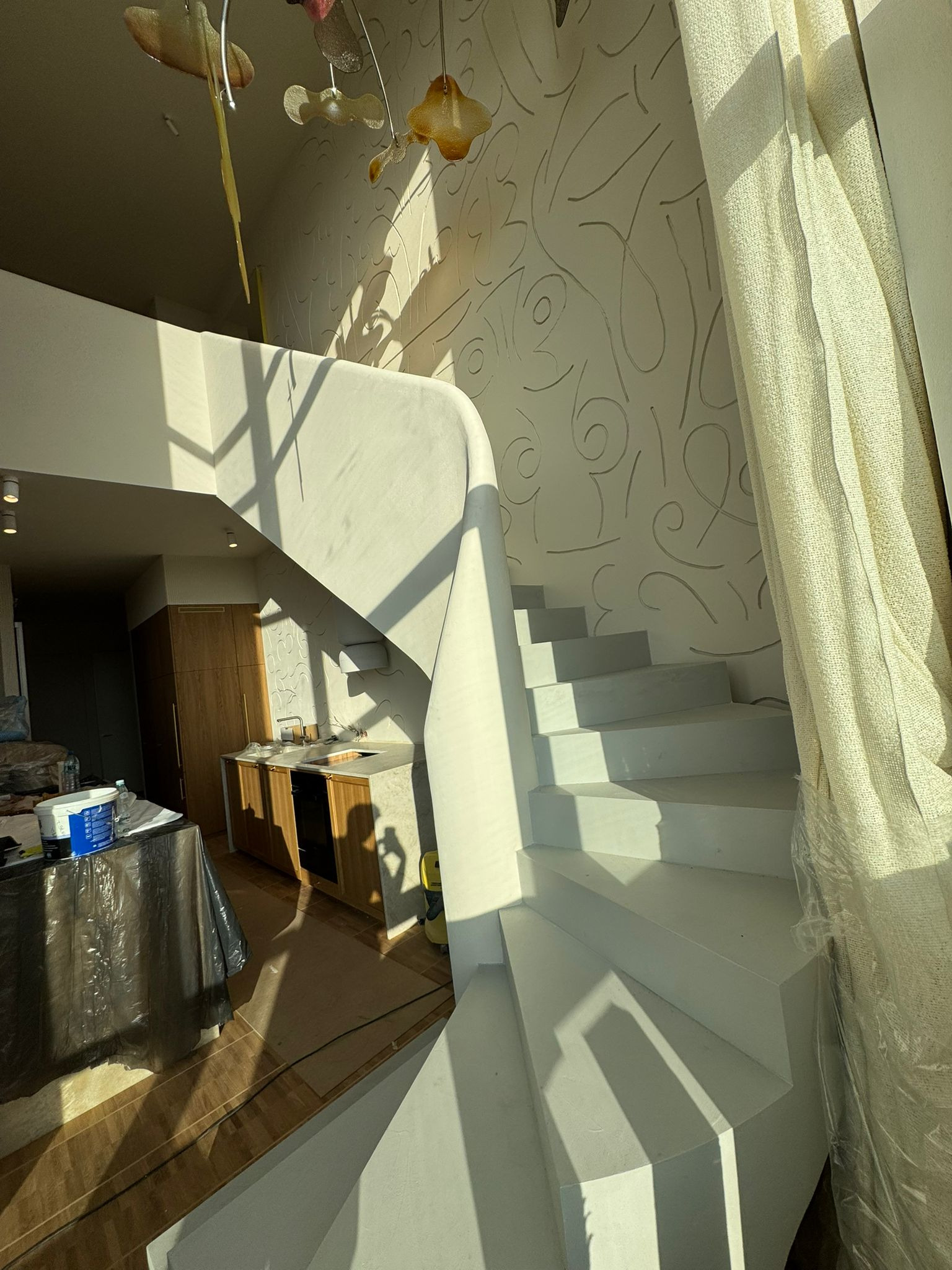 Białe schody w jasnym wnętrzu z dekoracyjną ścianą i zasłoniętym oknem. Widoczny fragment kuchni i lampa wisząca. Świeże malowanie, wpadające promienie słoneczne.