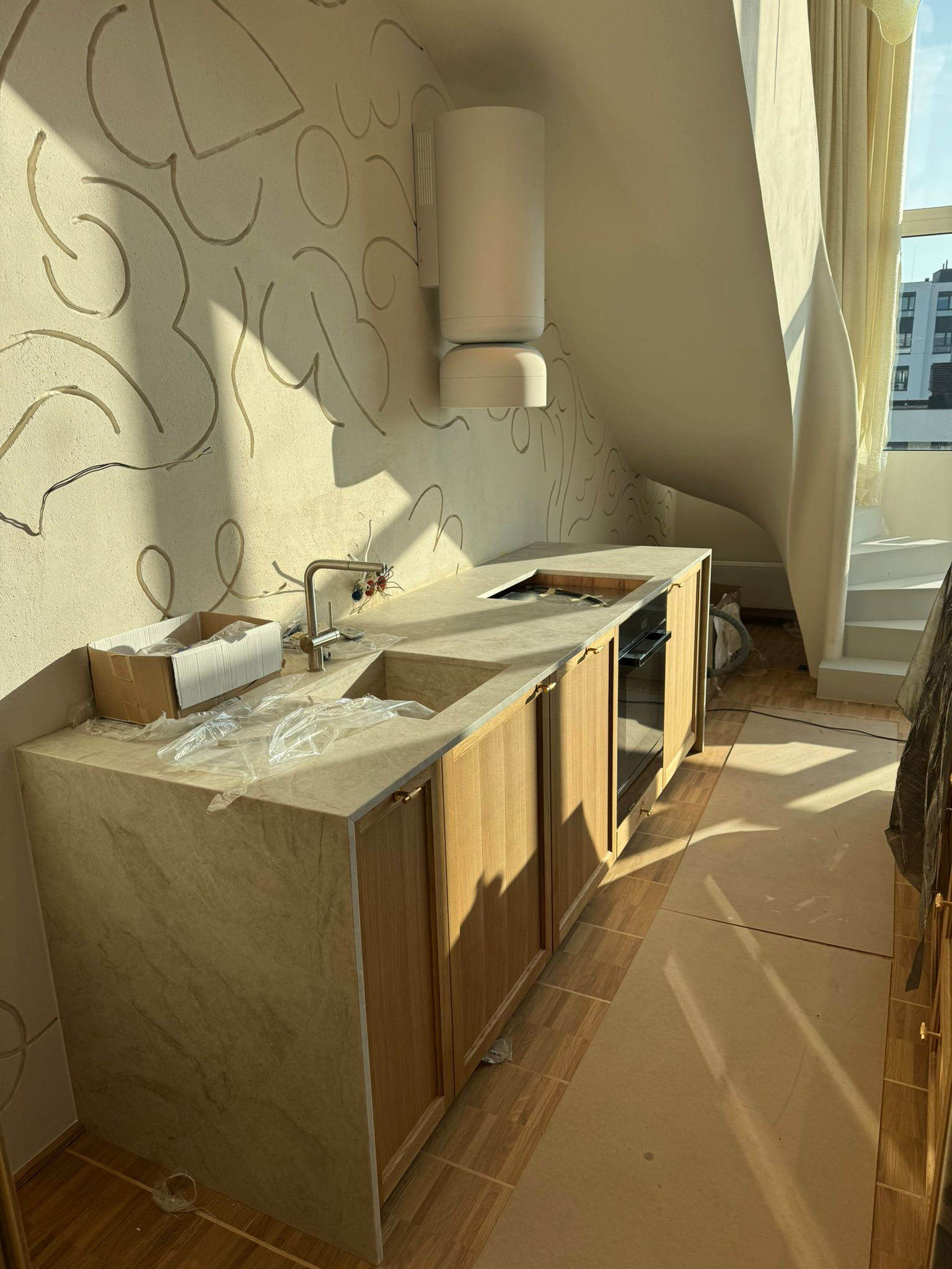 Jasna kuchnia w trakcie montażu w poddaszu ze skośnym sufitem, widoczne elementy instalacji i karton z akcesoriami. Na ścianie dekoracyjne, abstrakcyjne wzory.