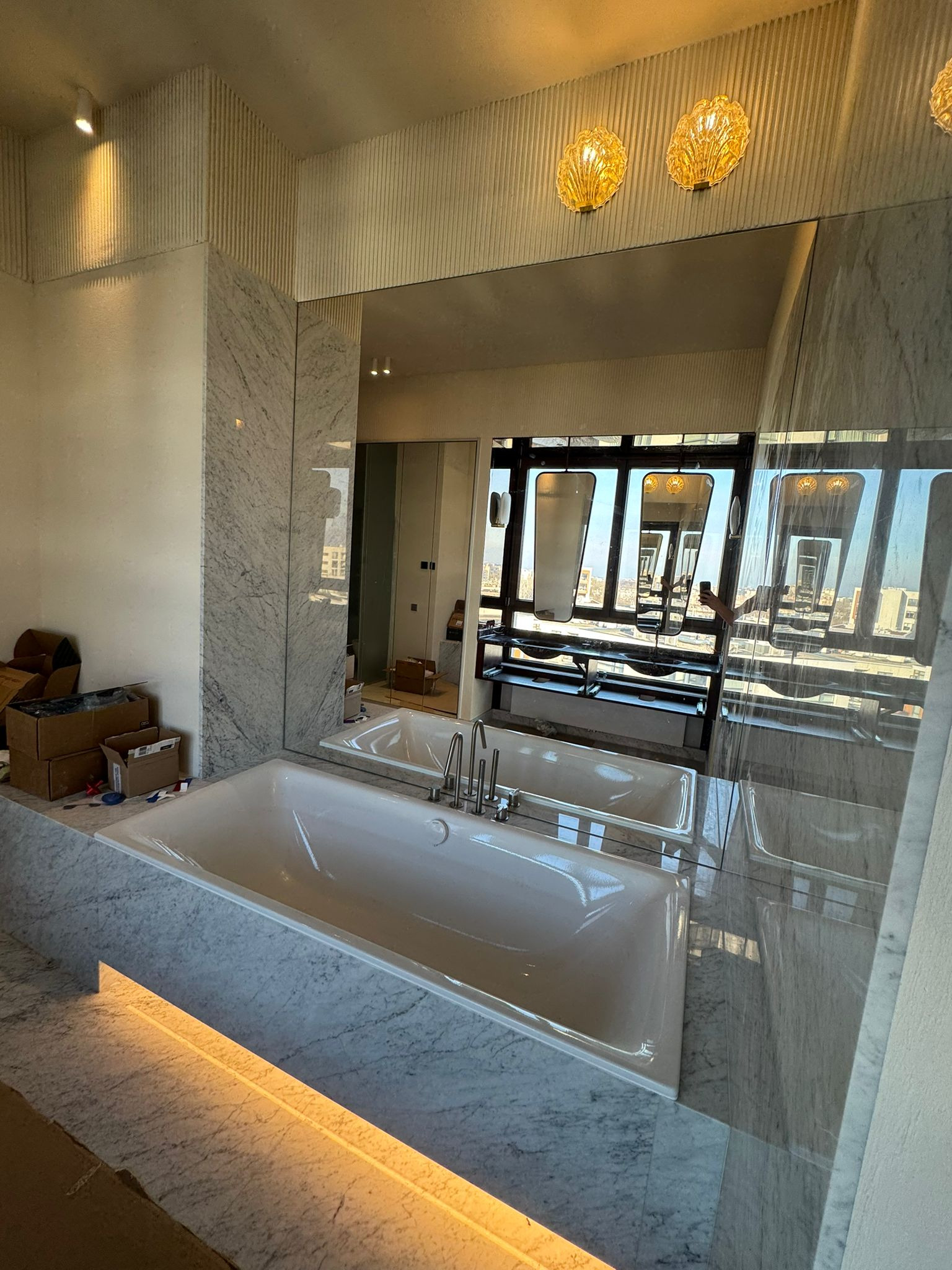 Nowoczesna łazienka z marmurowym wykończeniem, białą wanną i dużym lustrem odbijającym widok na miasto. Dekoracyjne oświetlenie ścienne.