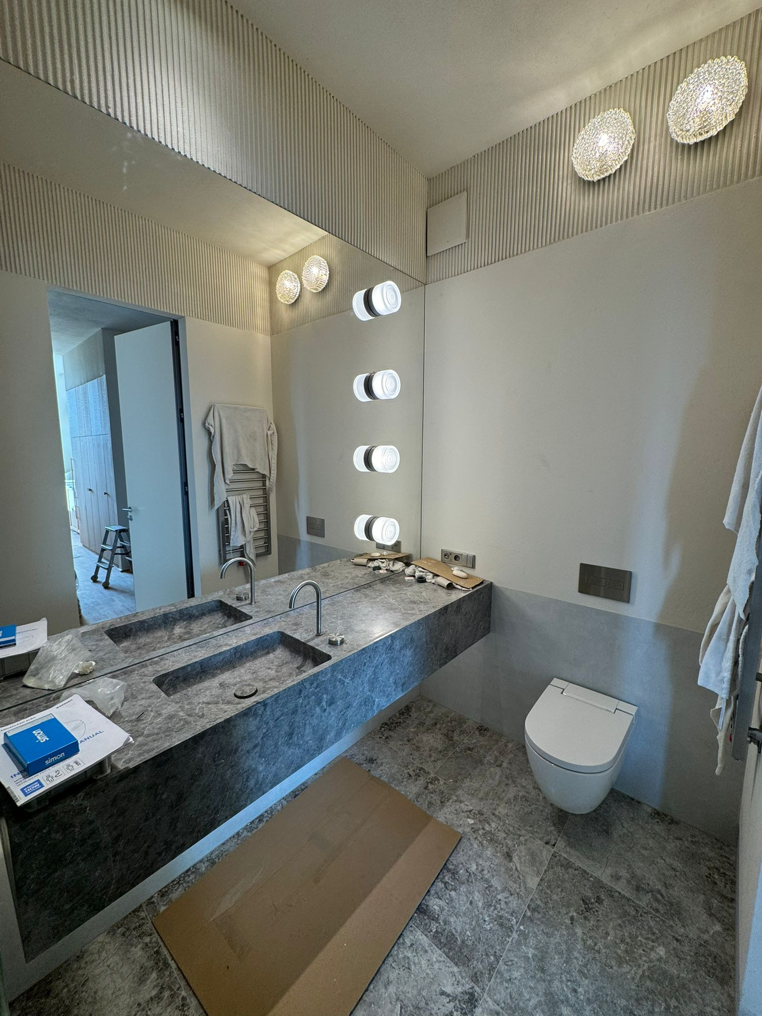 Nowoczesna łazienka w szarościach z marmurową umywalką i podświetlanym lustrem. Widoczne elementy wykończeniowe i karton na podłodze.