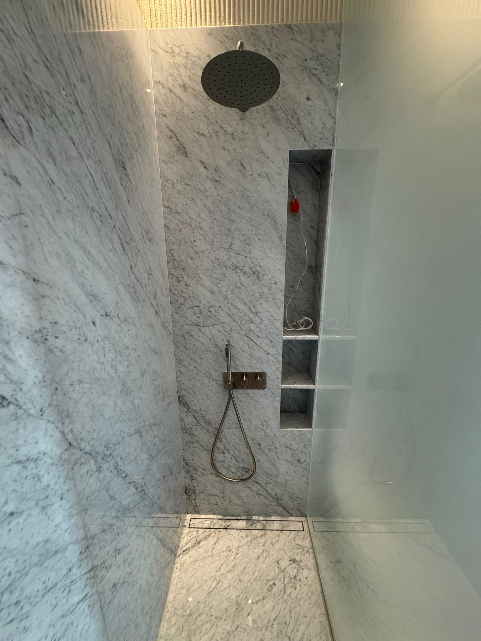 Nowoczesna kabina prysznicowa z marmurowymi ścianami, deszczownicą i wnęką z półkami. Minimalistyczny design, szklana ściana, odpływ liniowy. Eleganckie wykończenie łazienki.