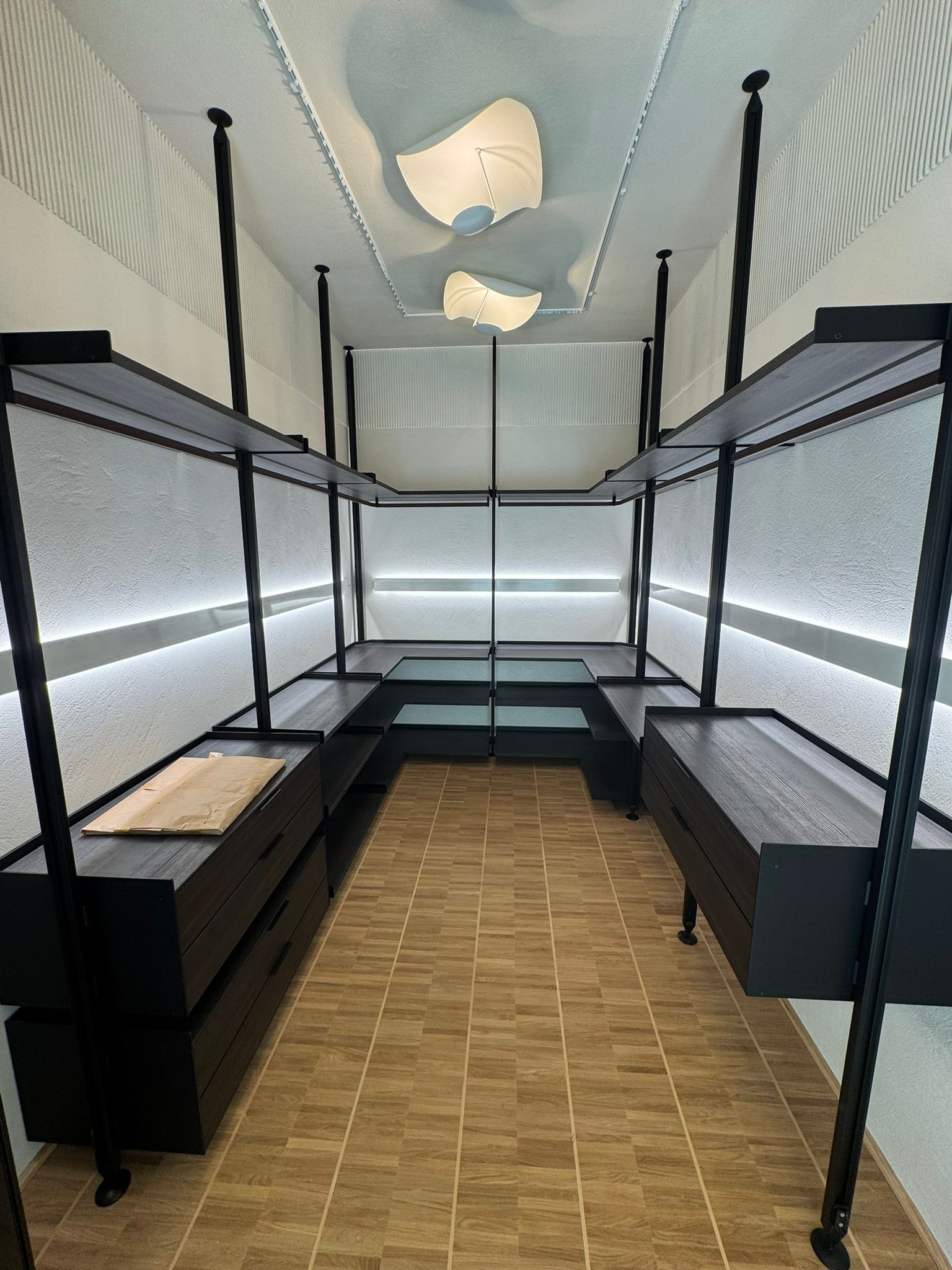 Nowoczesna garderoba z czarnymi regałami i szufladami, oświetleniem LED i beżową podłogą. Na półce leży brązowa paczka. Minimalistyczny design.