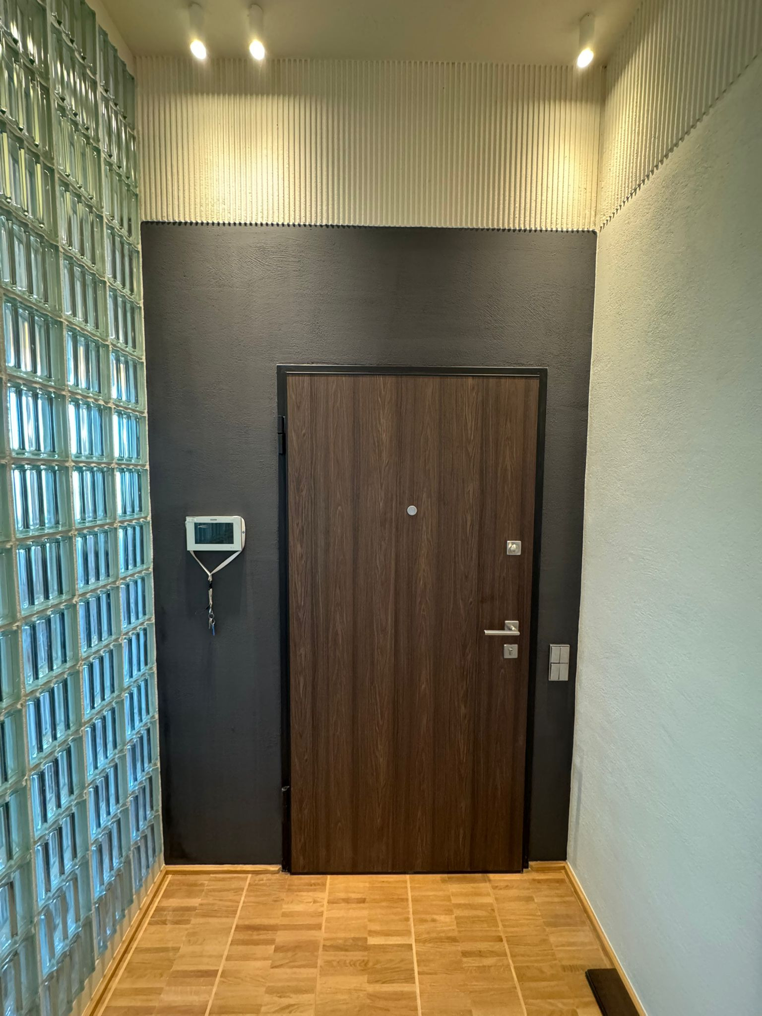 Nowoczesny przedpokój z ścianą z luksferów, drewnianymi panelami na suficie i ciemną ścianą z drewnianymi drzwiami. Podłoga wyłożona płytkami imitującymi drewno.