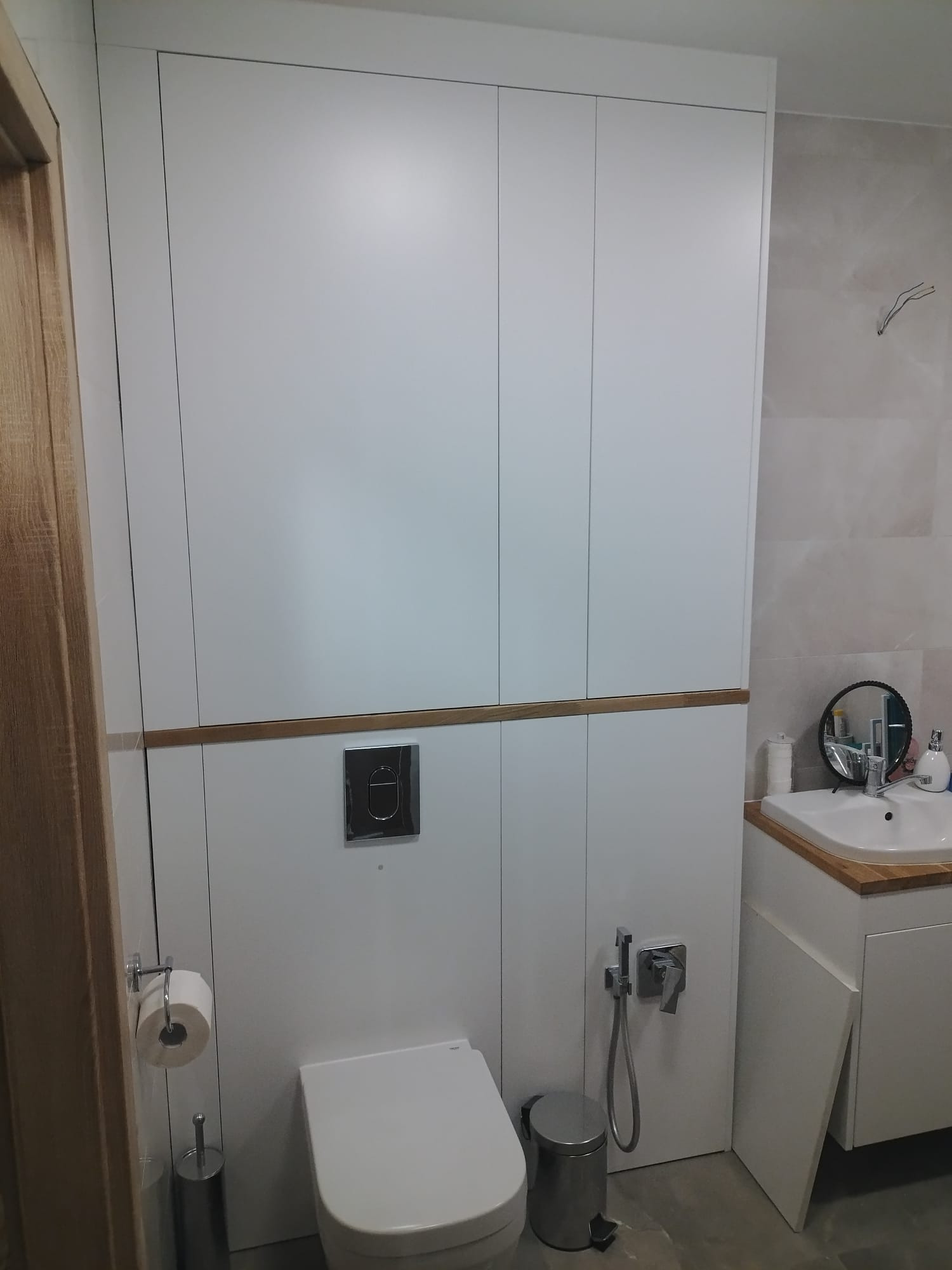 Nowoczesna łazienka z zabudową meblową w bieli, akcentowana drewnianym dekorem. Wisząca toaleta, bidetka i umywalka z okrągłym lustrem dopełniają minimalistyczny design.