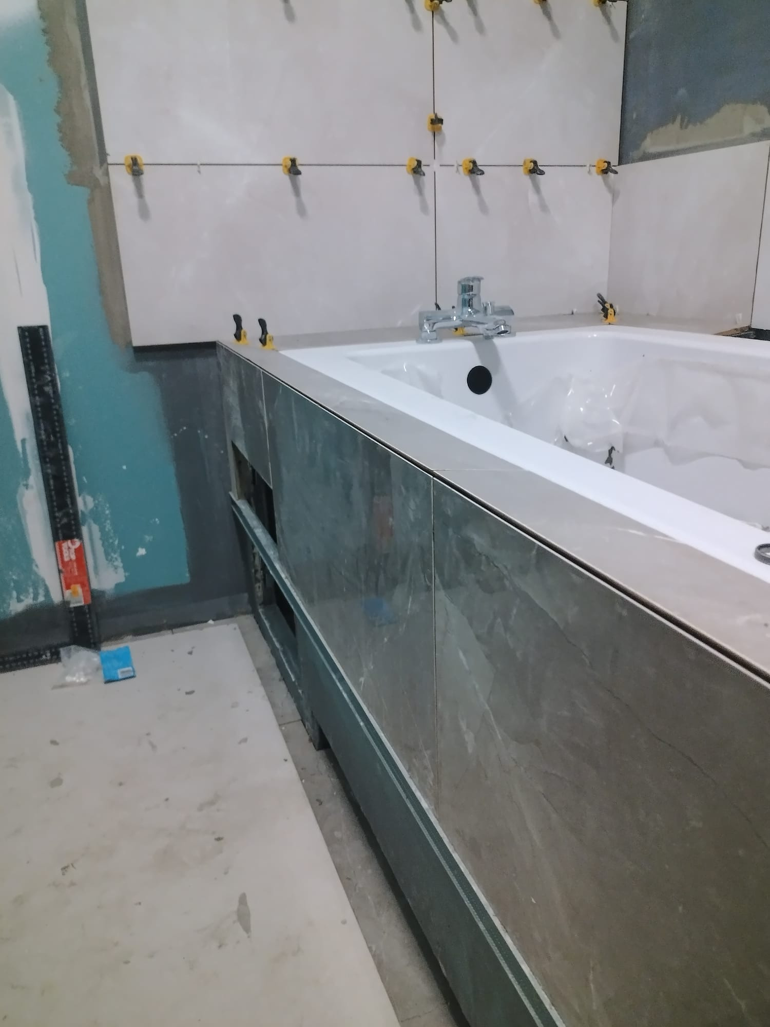 Wykończenie łazienki w trakcie: białe płytki na ścianie z żółtymi krzyżykami dystansowymi, obudowa wanny z szarych płytek imitujących marmur, widoczne narzędzia.