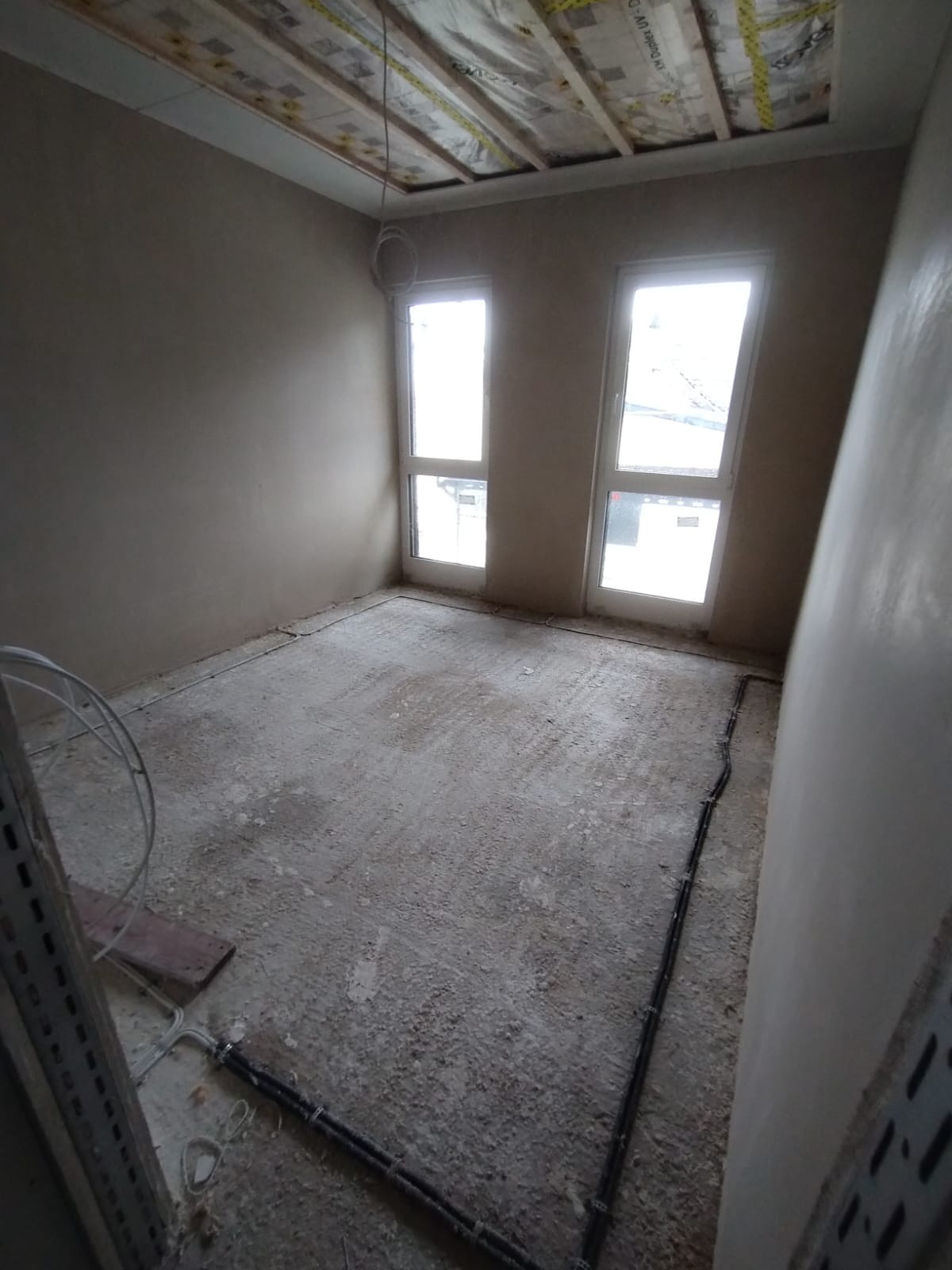 Pokój po nałożeniu tynku, widoczne dwa okna, instalacja elektryczna przy podłodze i sufit z izolacją. Ściany w kolorze beżowym, sufit biały.